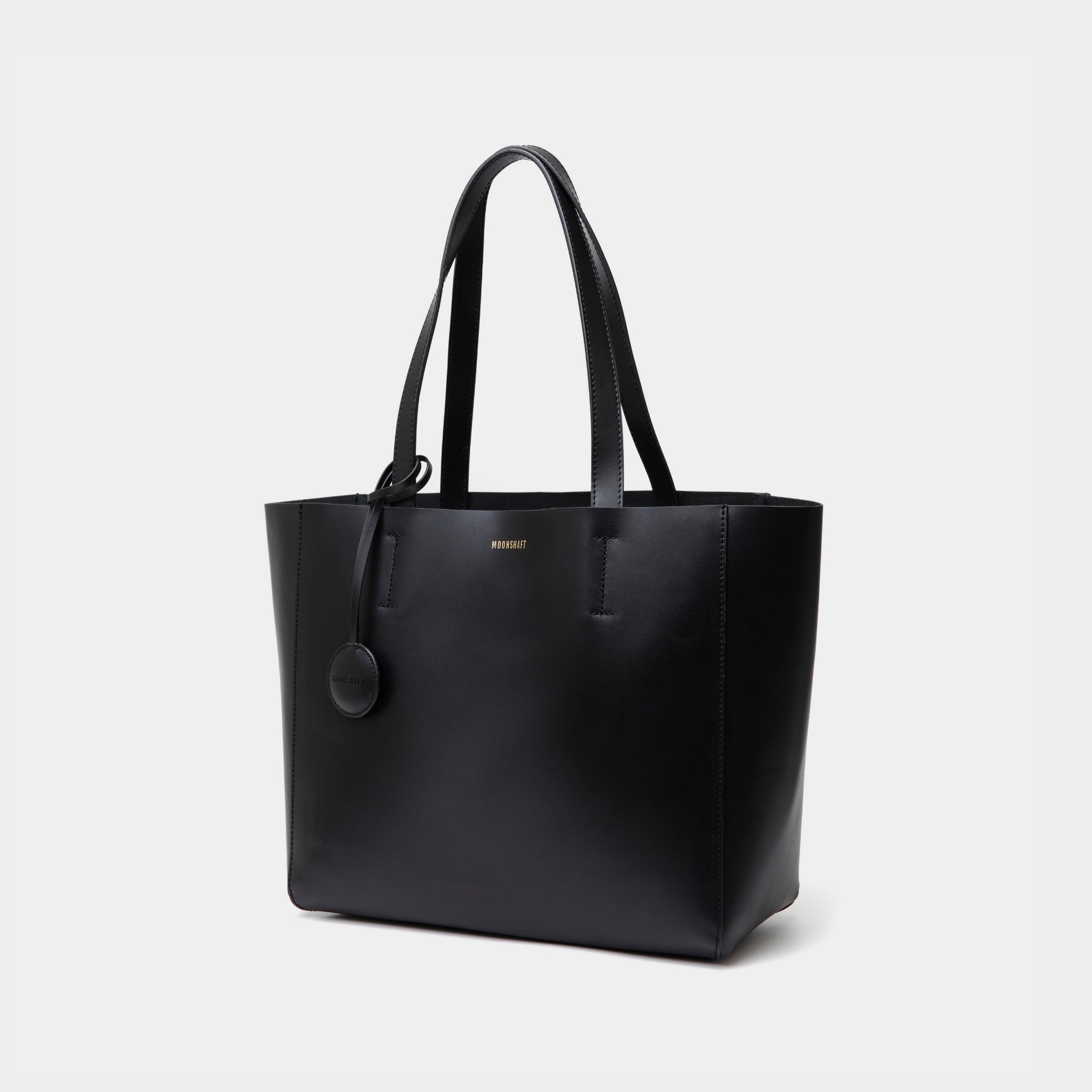 The diagonal view of the black MOONSHAFT tote bag displayed against a white background. The bag features a sleek, minimalist design with reinforced stitching along the handles, emphasizing its spacious and structured silhouette.