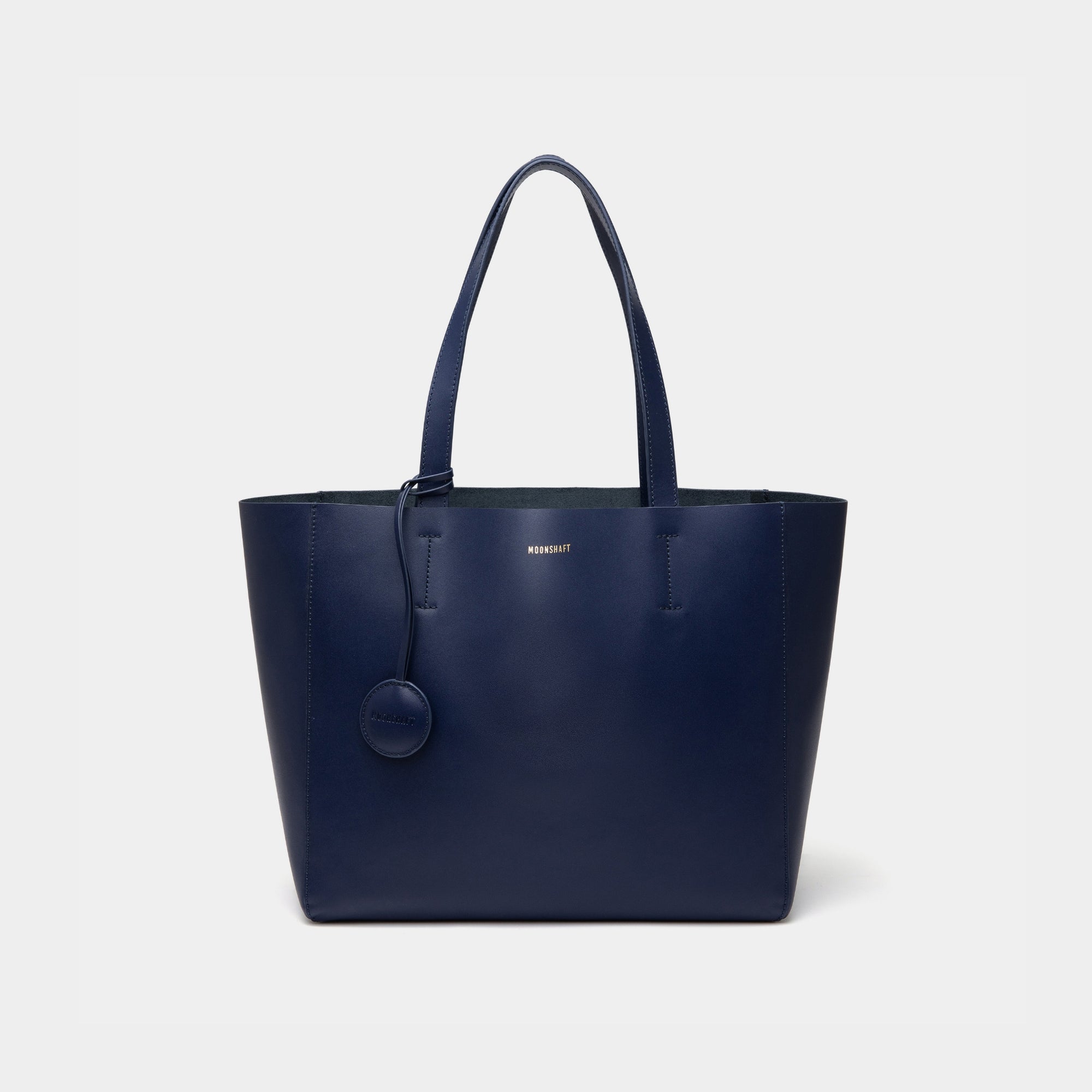A minimalist navy leather tote bag displayed against a plain background. The tote features a smooth leather texture and a clean, structured design with no external embellishments, showcasing its simplicity and elegance. It has two sturdy leather handles and a spacious interior, making it a practical choice for everyday use. The neutral tan color adds to the bag’s versatile appeal, perfect for complementing various outfits and occasions.