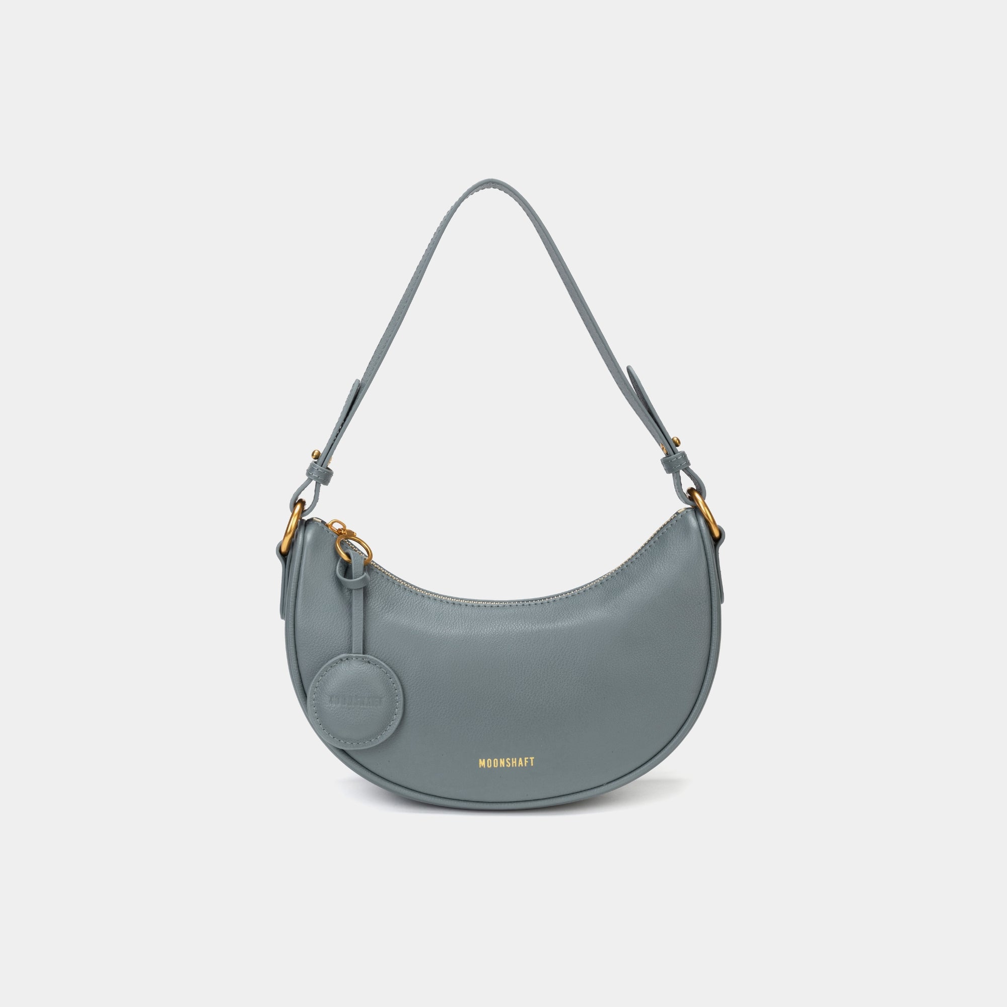 Front view of the Crescent Moon bag in ash blue, showcasing its smooth leather finish, curved silhouette, and gold hardware. The top handle is visible, along with an attached round coin purse accessory on the side. The minimal and elegant design highlights the bag's clean lines and structured shape.