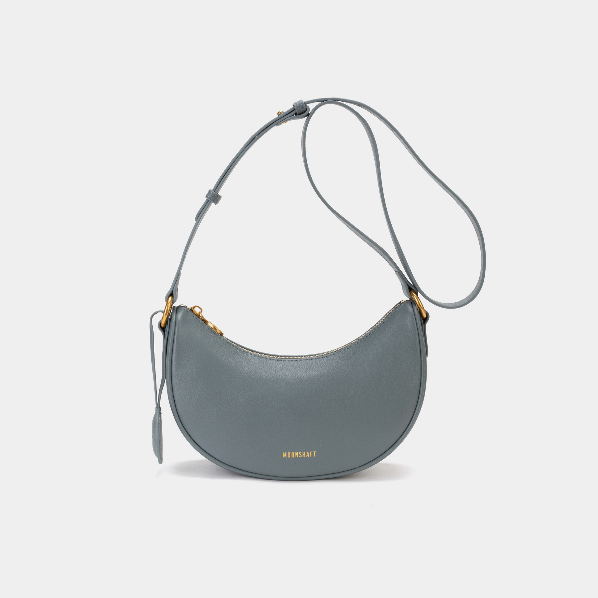 Front view of the Crescent Moon bag in ash blue, featuring smooth leather with a minimalist design. The bag is shown with its adjustable crossbody strap and gold-tone hardware, highlighting the curved silhouette and simple elegance of the design.