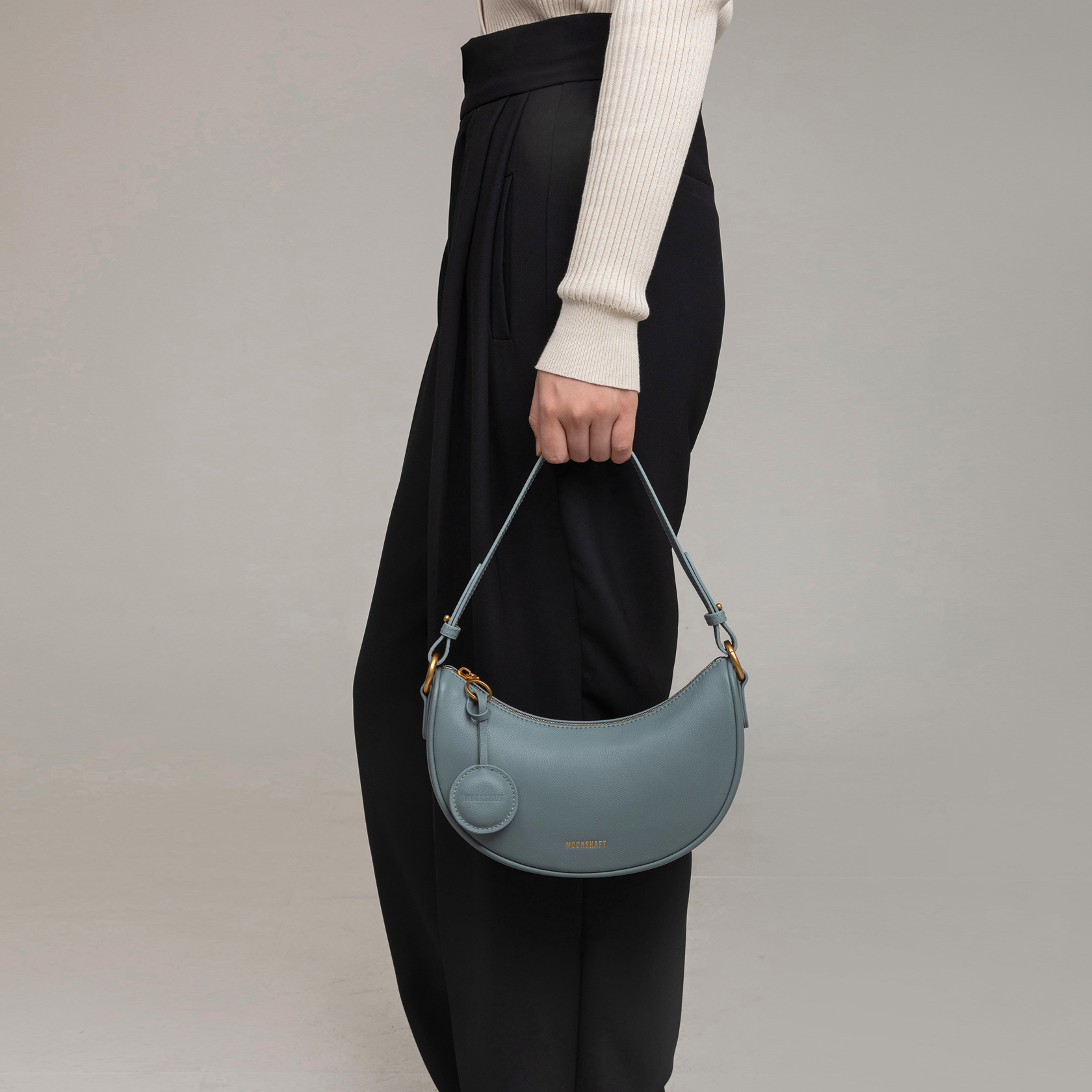 Close-up of a model holding the Crescent Moon bag in ash blue smooth leather by her side. The bag is carried by its short handle, showcasing its compact and curved shape. The model wears black high-waisted trousers with pleats, paired with a beige ribbed long-sleeve top, complementing the bag's sophisticated and minimalist design.