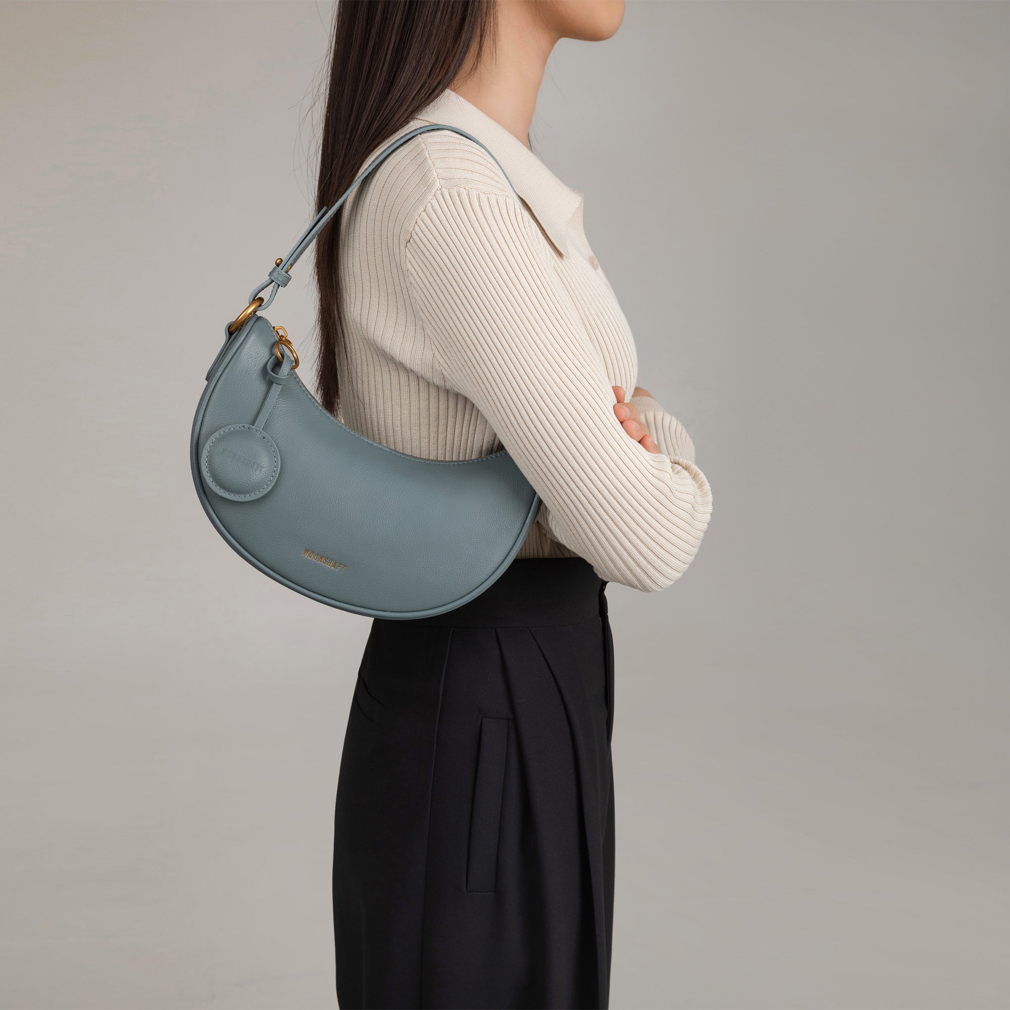Model wearing the Crescent Moon bag in ash blue smooth leather over her shoulder. The bag is styled with a beige ribbed top and black pleated trousers, showing its versatility and chic design. The sleek, curved silhouette of the bag sits comfortably under the arm, complemented by its minimalistic details and gold hardware.