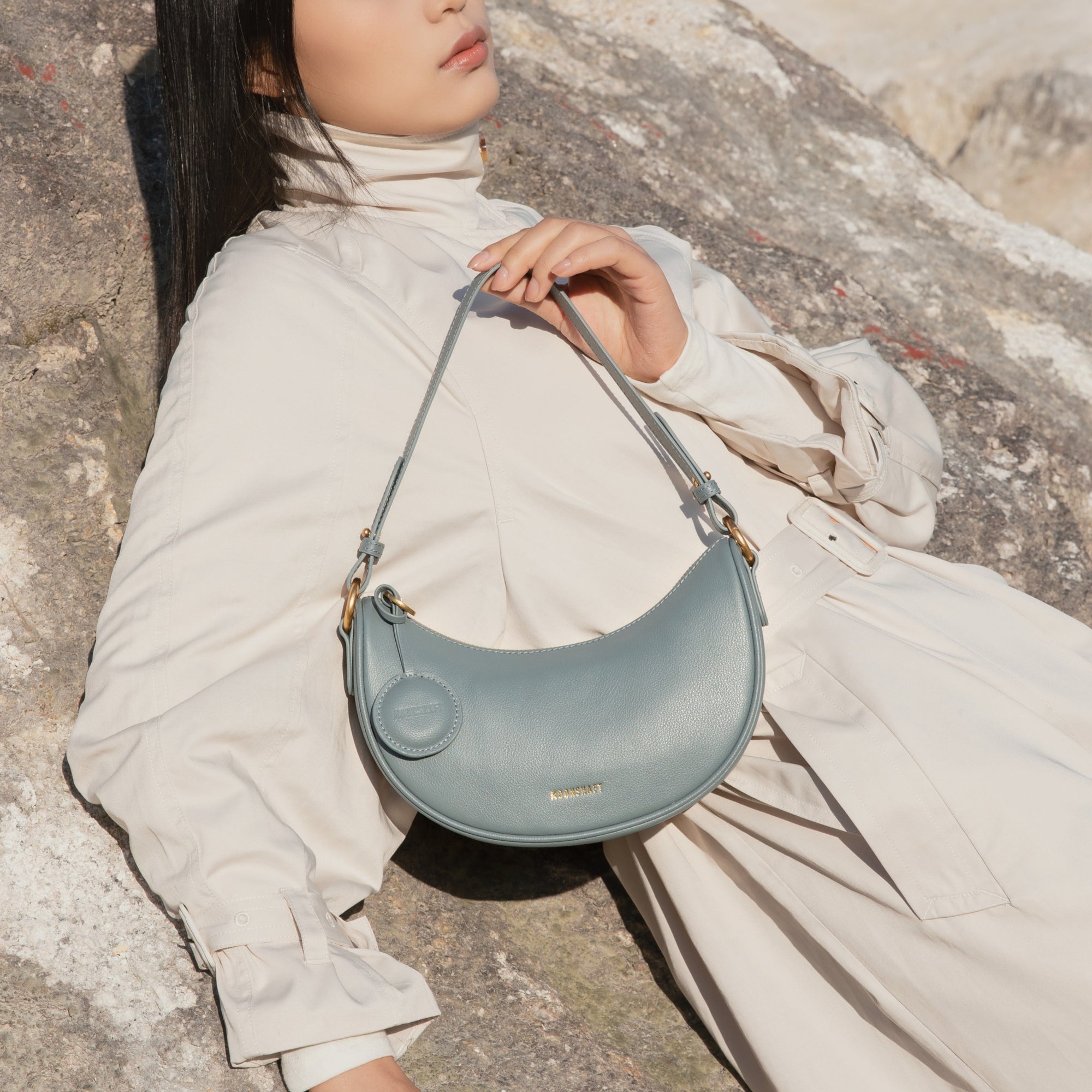 A model dressed in a light beige outfit lounges against a stone backdrop, holding the Crescent Moon bag in ash blue. The smooth leather bag, with its gold-tone hardware and attached coin pouch, rests elegantly on her lap. The bag's soft, crescent shape complements the minimalist and relaxed styling, while the MOONSHAFT logo in gold adds a touch of luxury. The natural outdoor setting, combined with the muted tones of the model's attire and the bag, creates a serene and sophisticated look.