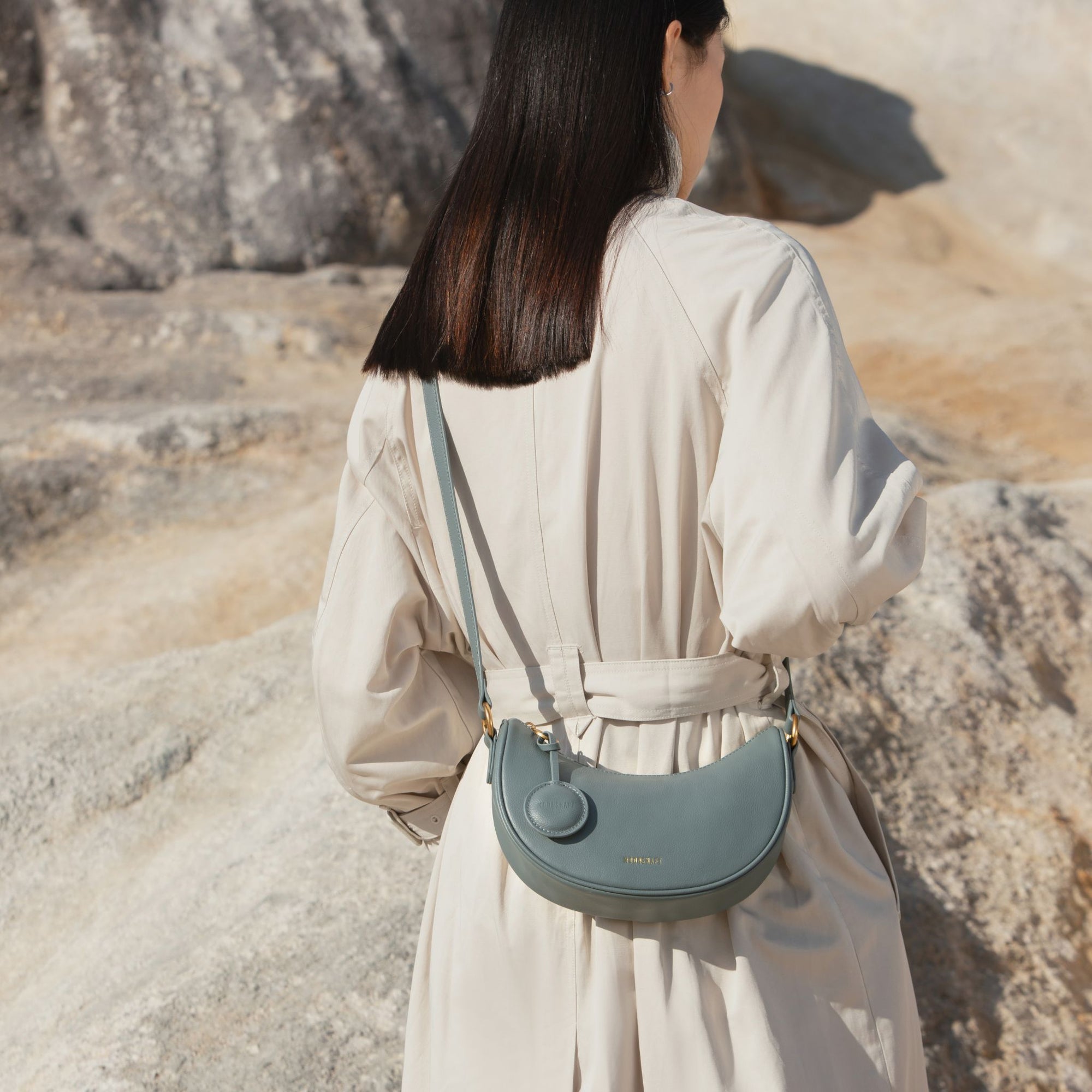 Back view of a model wearing a light beige trench coat and carrying the Crescent Moon bag in ash blue. The smooth leather bag, featuring gold-tone hardware and a coin pouch attached to the side, hangs crossbody with the adjustable strap. The bag’s soft, rounded shape contrasts elegantly with the neutral tones of the coat and the rugged outdoor stone backdrop. The MOONSHAFT logo in gold is subtly visible on the back of the bag, complementing its minimal and sophisticated design.