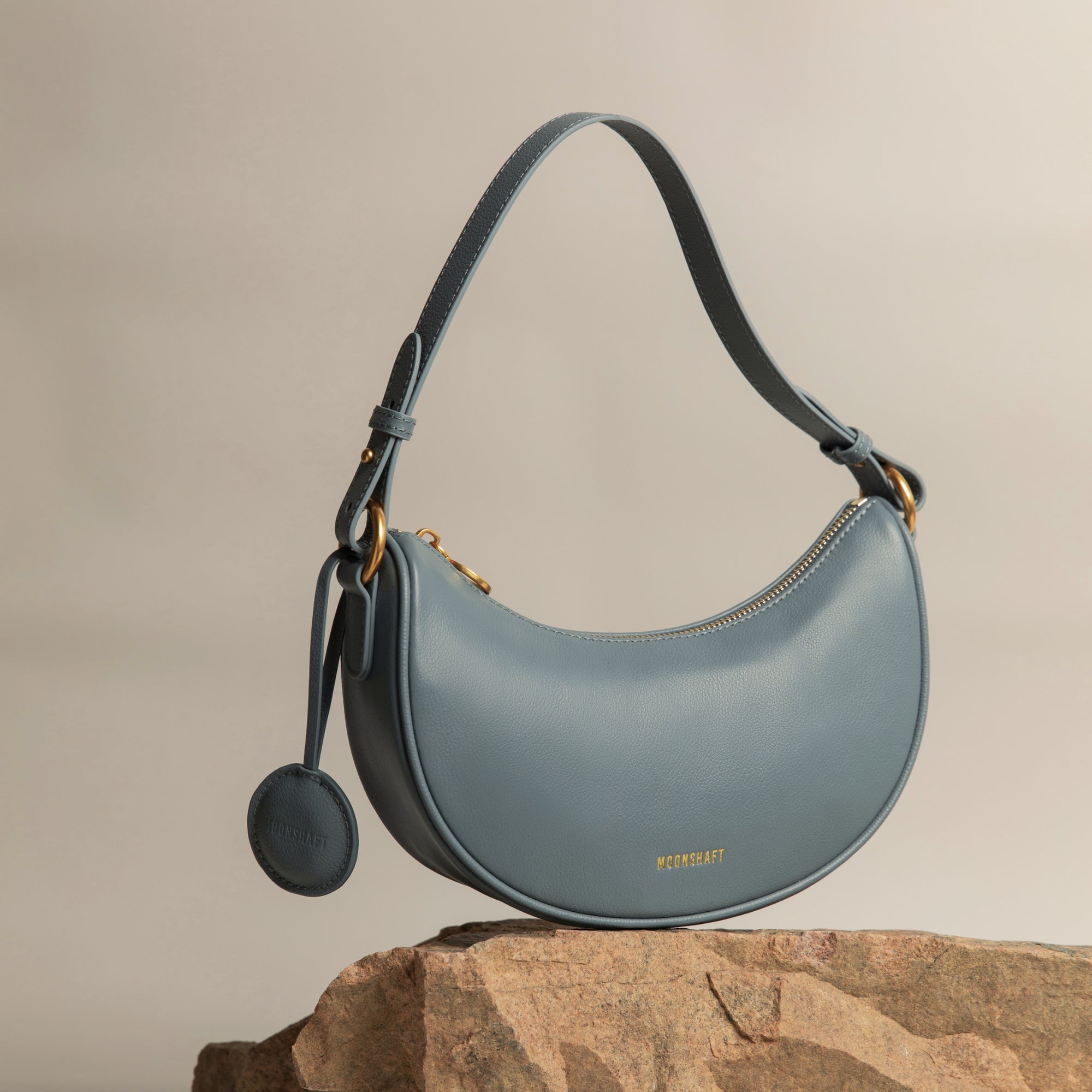 Crescent Moon bag in ash blue smooth leather, elegantly displayed on a stone surface with gold hardware details. The bag's curved silhouette and minimalist design are complemented by a detachable leather tag, set against a neutral-toned background, highlighting its premium craftsmanship and modern aesthetic.