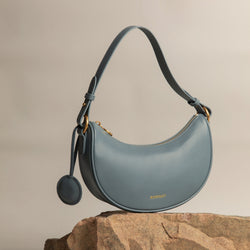Crescent Moon bag in ash blue smooth leather, elegantly displayed on a stone surface with gold hardware details. The bag's curved silhouette and minimalist design are complemented by a detachable leather tag, set against a neutral-toned background, highlighting its premium craftsmanship and modern aesthetic.