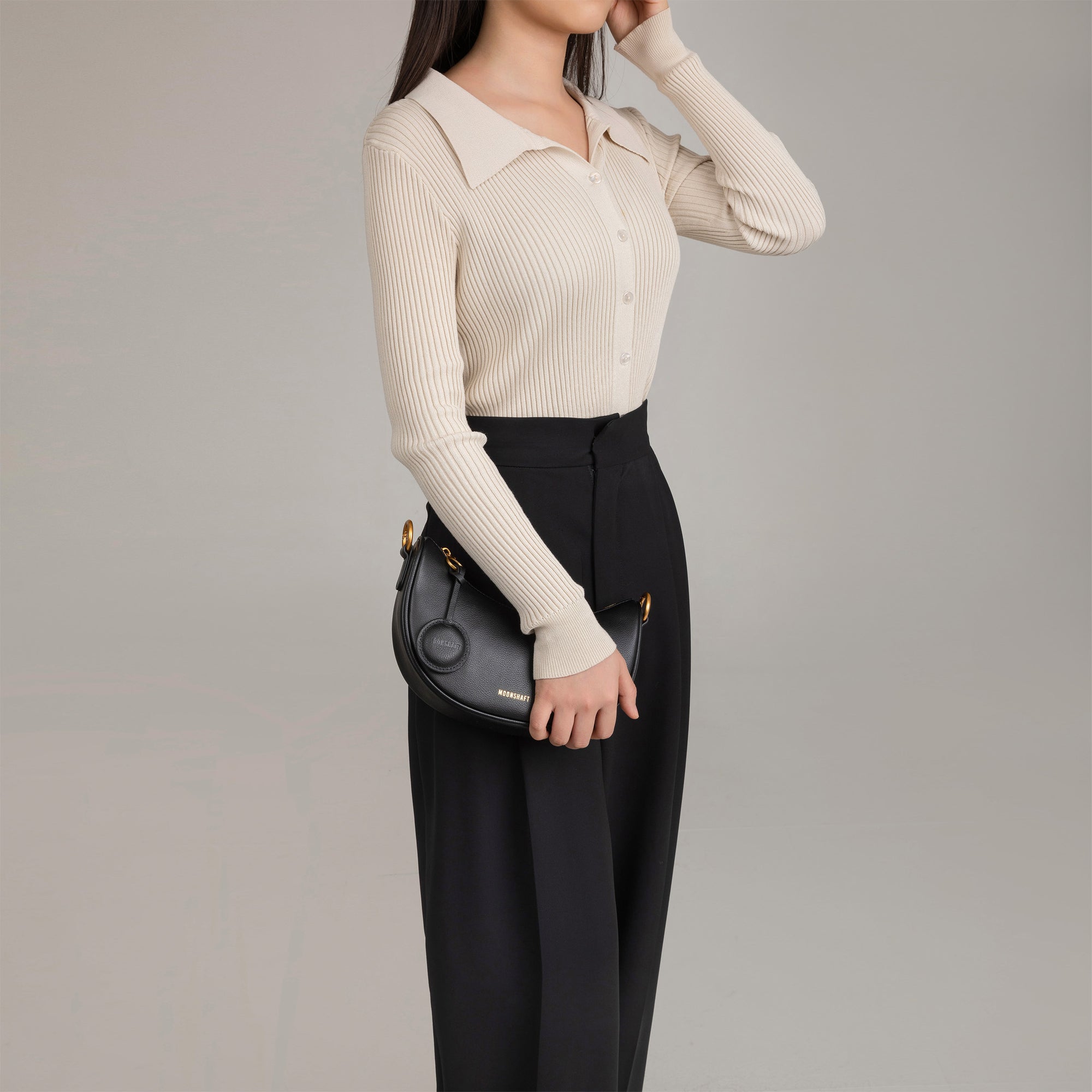Model holding the Crescent Moon bag in black smooth leather at her side, wearing a beige ribbed button-up top paired with black pleated trousers. The sleek, minimalist design of the bag is highlighted by its compact size, curved shape, and gold hardware, making it a versatile accessory for both casual and formal outfits.
