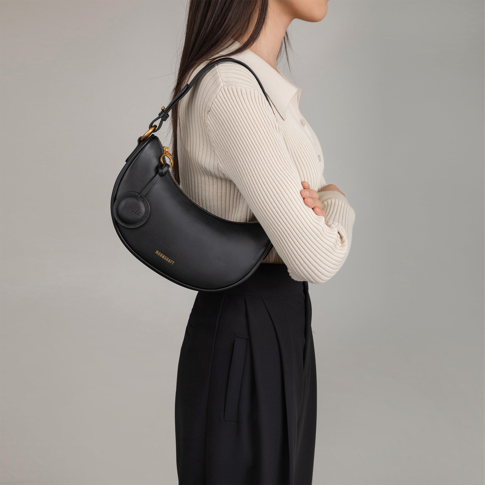 Model wearing the Crescent Moon bag in black smooth leather over her shoulder. The bag is styled with a beige ribbed top and black pleated trousers, showing its versatility and chic design. The sleek, curved silhouette of the bag sits comfortably under the arm, complemented by its minimalistic details and gold hardware.