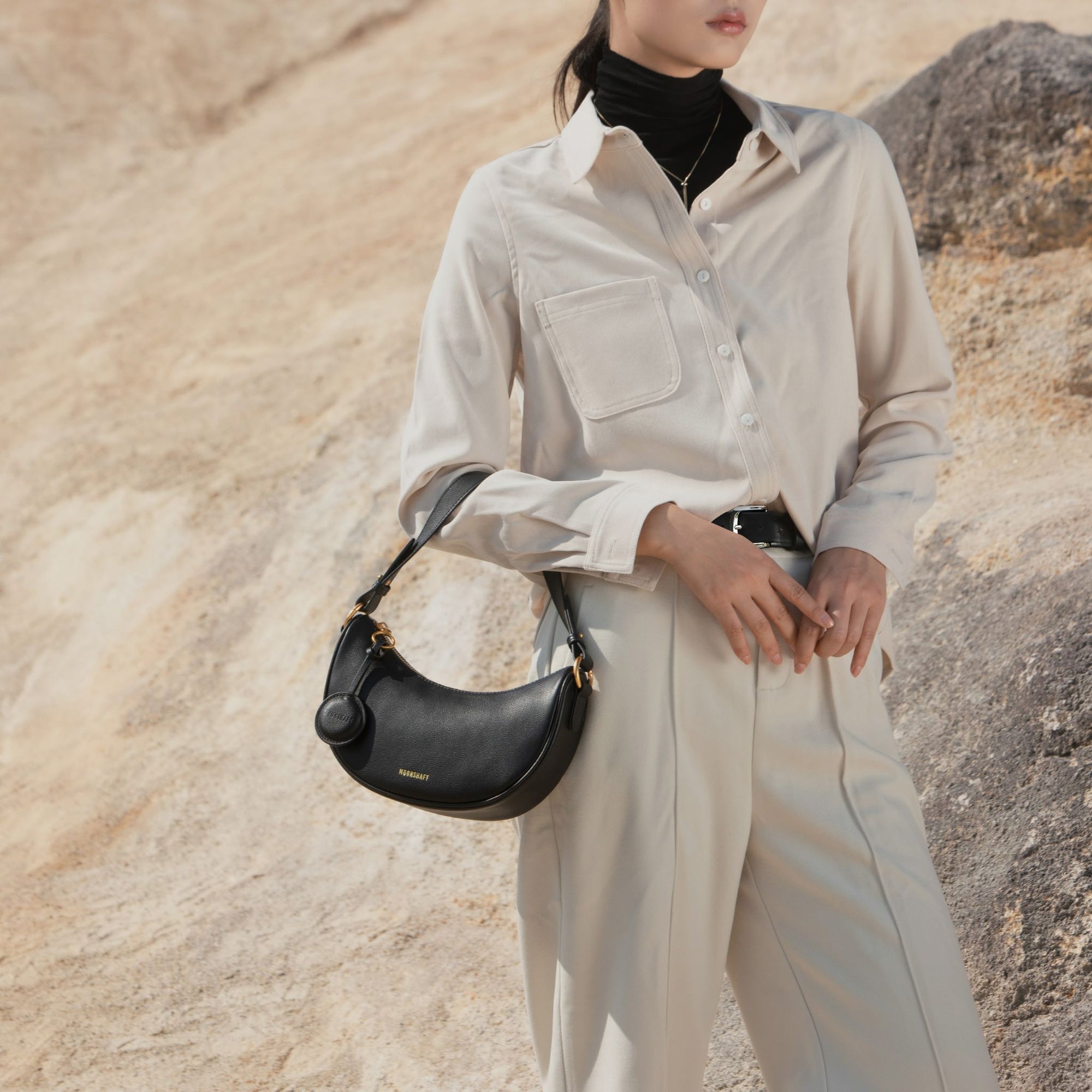 A model styled with the Crescent Moon bag in black, paired with a beige button-up shirt and neutral trousers. The smooth leather bag, featuring gold-tone details, is worn on the shoulder, complementing the minimal and elegant outfit.