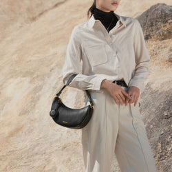 A model styled with the Crescent Moon bag in black, paired with a beige button-up shirt and neutral trousers. The smooth leather bag, featuring gold-tone details, is worn on the shoulder, complementing the minimal and elegant outfit.