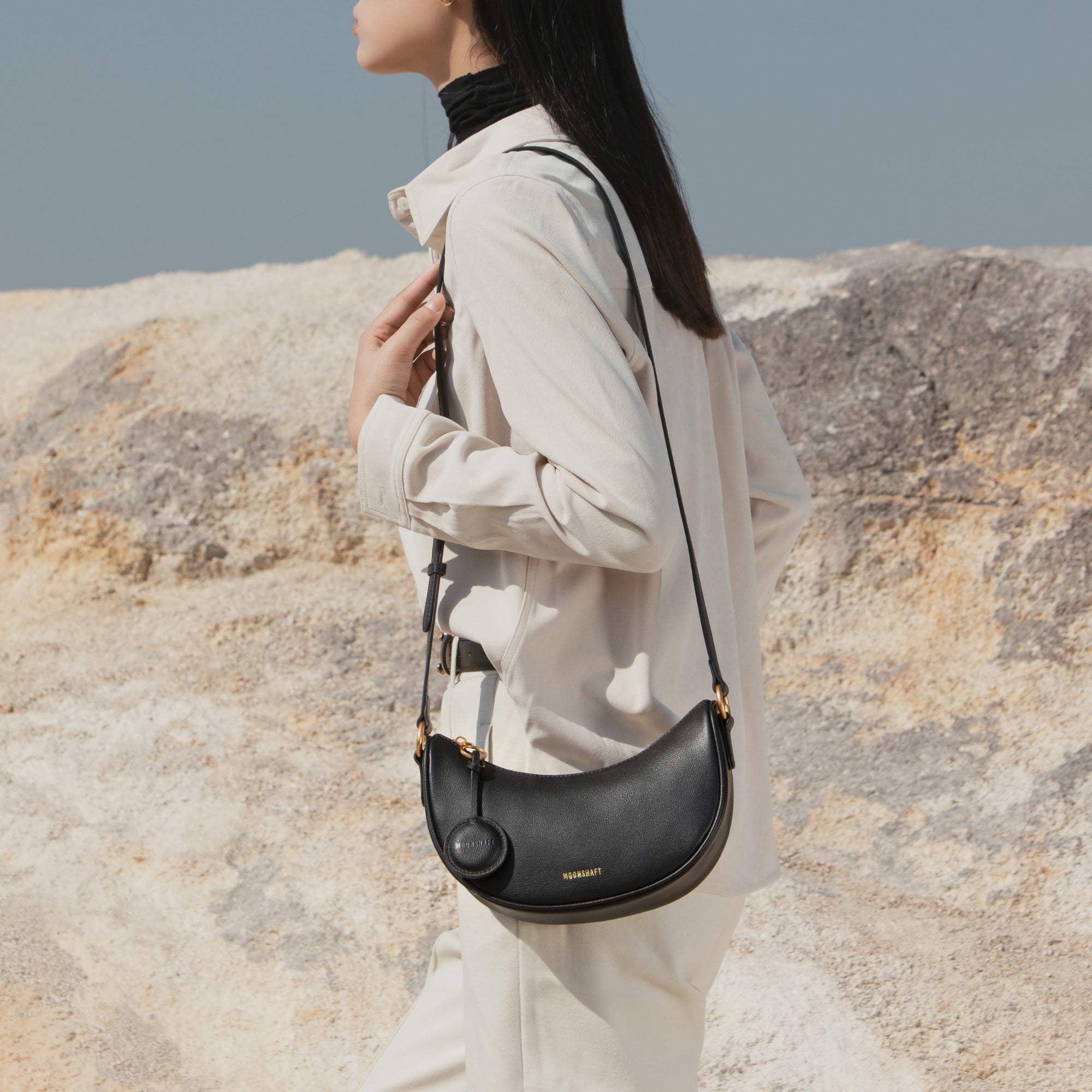A side profile of a model wearing the Crescent Moon bag in black, styled crossbody over a light beige shirt and trousers. The smooth leather bag adds a sleek touch to the neutral-toned outfit, perfect for a minimalist yet chic look.