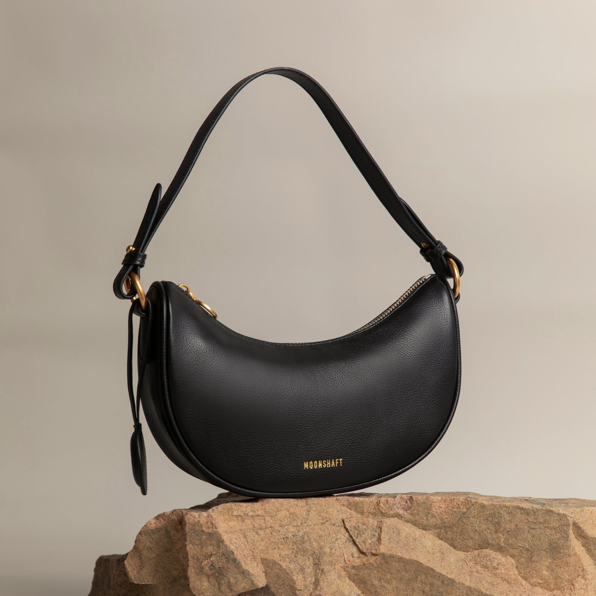 Crescent Moon bag in black smooth leather, elegantly displayed on a stone surface with gold hardware details. The bag's curved silhouette and minimalist design are complemented by a detachable leather tag, set against a neutral-toned background, highlighting its premium craftsmanship and modern aesthetic.