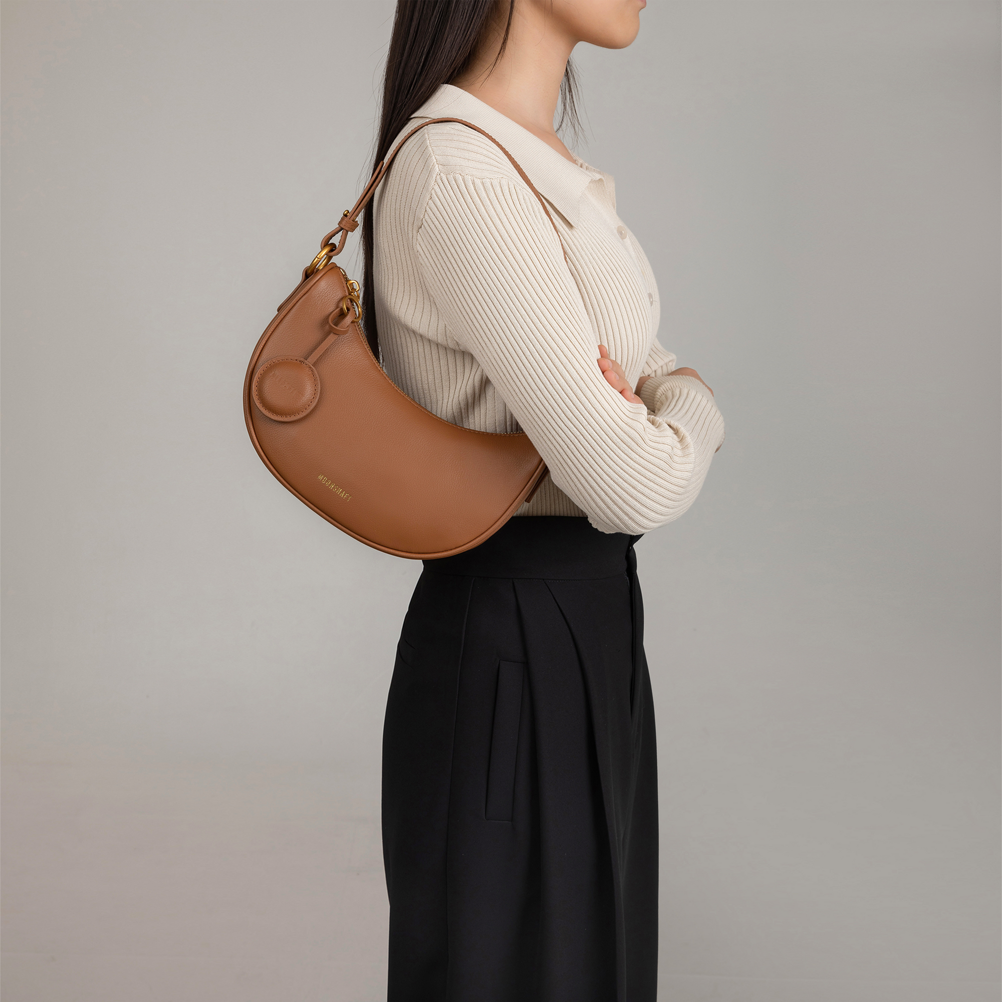 Model wearing the Crescent Moon bag in caramel smooth leather over her shoulder. The bag is styled with a beige ribbed top and black pleated trousers, showing its versatility and chic design. The sleek, curved silhouette of the bag sits comfortably under the arm, complemented by its minimalistic details and gold hardware.
