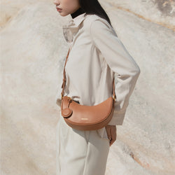 A woman wearing a neutral-toned outfit, carrying the Crescent Moon bag in caramel smooth leather across her body. The bag, styled as a crossbody, features a round leather tag and gold hardware, complementing her minimalist, natural look against an outdoor backdrop with soft, muted earthy tones.
