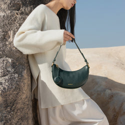 Model leaning against a textured rock in an outdoor setting, holding the Crescent Moon bag in pine grove green. The model is dressed in a soft, oversized cream sweater and light-colored pants, complementing the natural and earthy tones of the setting. The smooth leather bag, with its gold-toned hardware and minimalist design, stands out against the serene, neutral background, adding an element of sophistication to the relaxed, natural aesthetic.