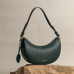 Crescent Moon bag in a deep pine grove green color, resting elegantly on a textured stone. The bag features smooth leather, gold-toned hardware, and a round leather charm hanging from the side. Its curved shape and minimalist design are complemented by the soft neutral background, highlighting the rich, luxurious tone of the bag.