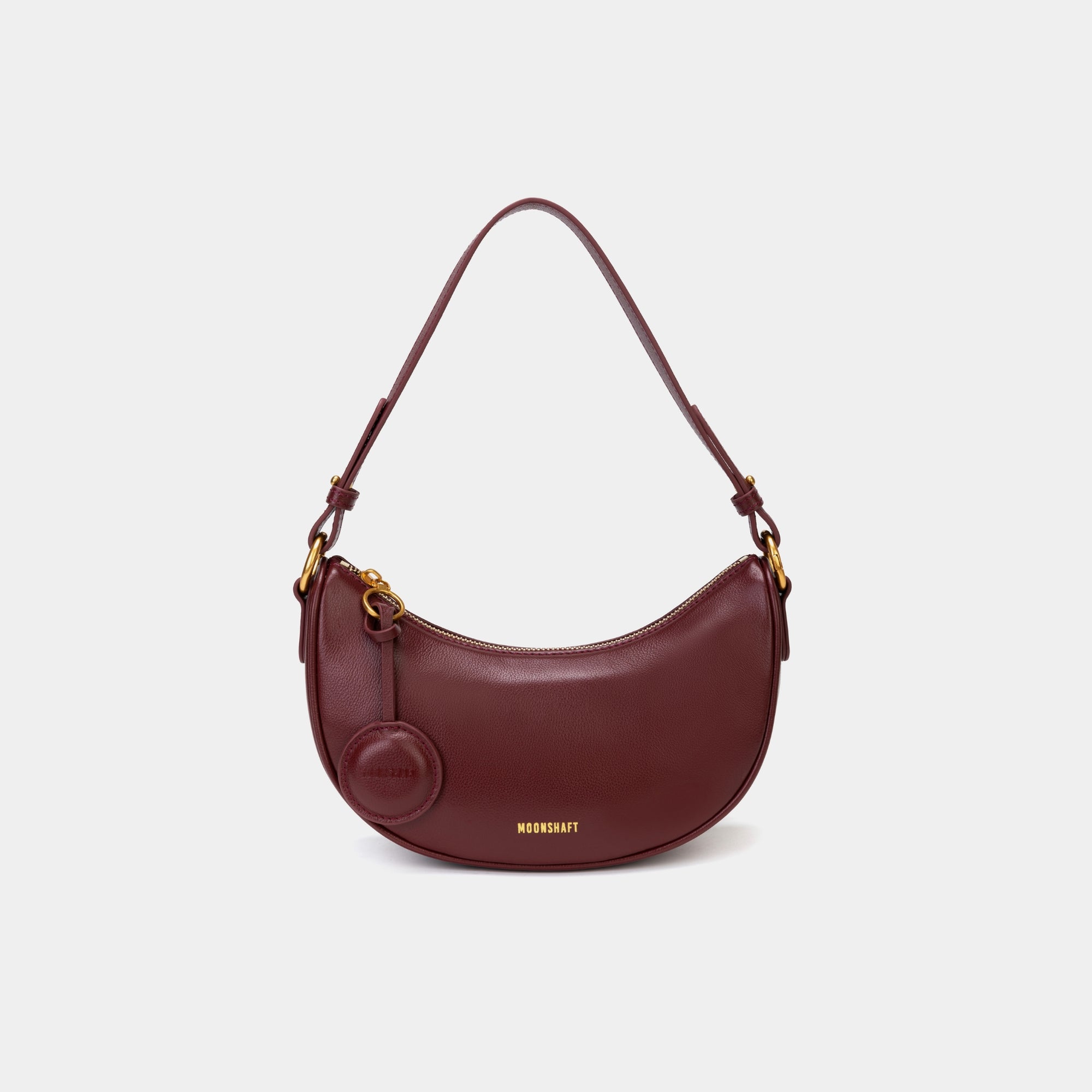 Front view of the Crescent Moon bag in wine, showcasing its smooth leather finish, curved silhouette, and gold hardware. The top handle is visible, along with an attached round coin purse accessory on the side. The minimal and elegant design highlights the bag's clean lines and structured shape.