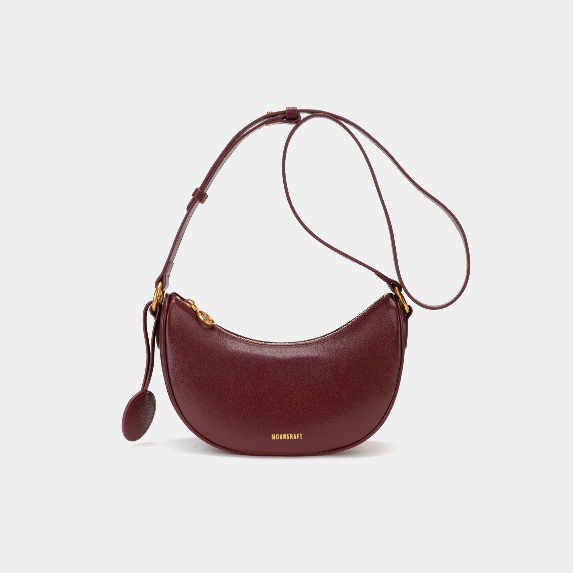 Front view of the Crescent Moon bag in wine, featuring smooth leather with a minimalist design. The bag is shown with its adjustable crossbody strap and gold-tone hardware, highlighting the curved silhouette and simple elegance of the design.