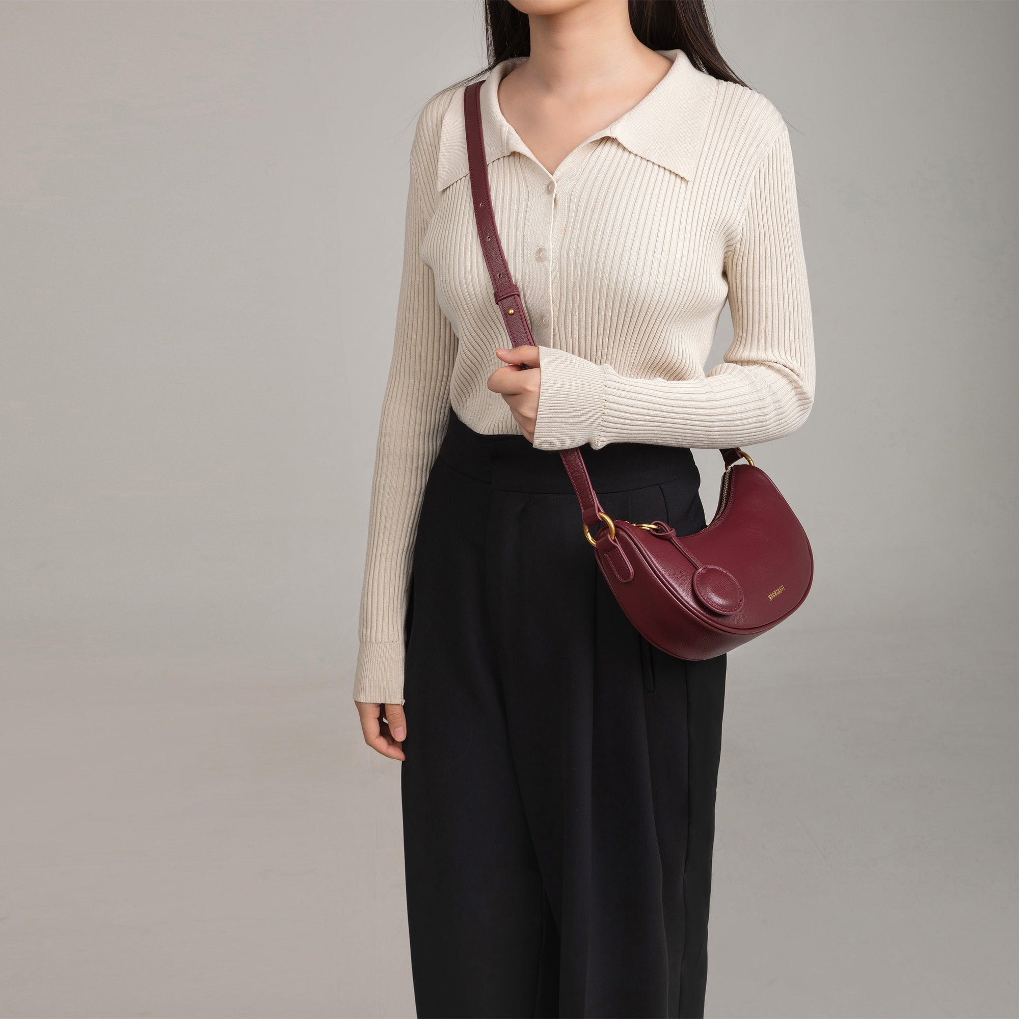 Model wearing the Crescent Moon bag in wine smooth leather across her shoulder. The bag is worn with a longer strap, draped casually at the hip. The model wears a beige ribbed long-sleeve top with a collar, paired with black high-waisted pleated trousers, creating a chic and elegant look.