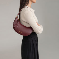 Model wearing the Crescent Moon bag in wine smooth leather over her shoulder. The bag is styled with a beige ribbed top and black pleated trousers, showing its versatility and chic design. The sleek, curved silhouette of the bag sits comfortably under the arm, complemented by its minimalistic details and gold hardware.