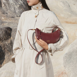 A model holding the Crescent Moon bag in wine, styled with a beige trench coat. The smooth leather bag features gold-tone details and is carried in hand for an elegant, minimalistic look.