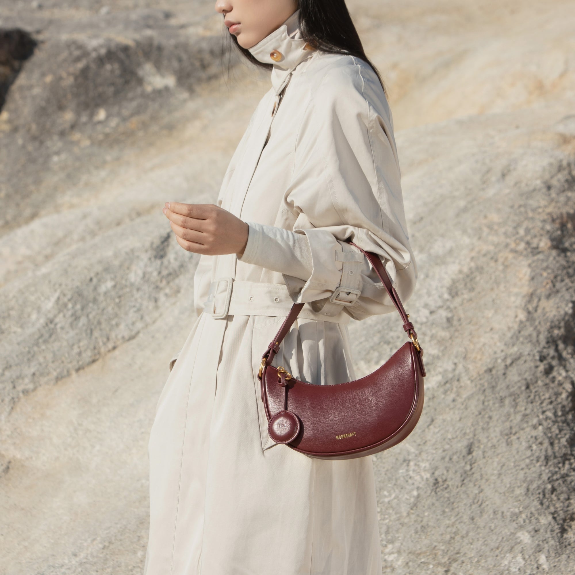 A model outdoors wearing a beige trench coat, carrying the Crescent Moon bag in wine. The smooth leather bag with gold-tone hardware is worn on the shoulder, adding a refined touch to the minimalist outfit.