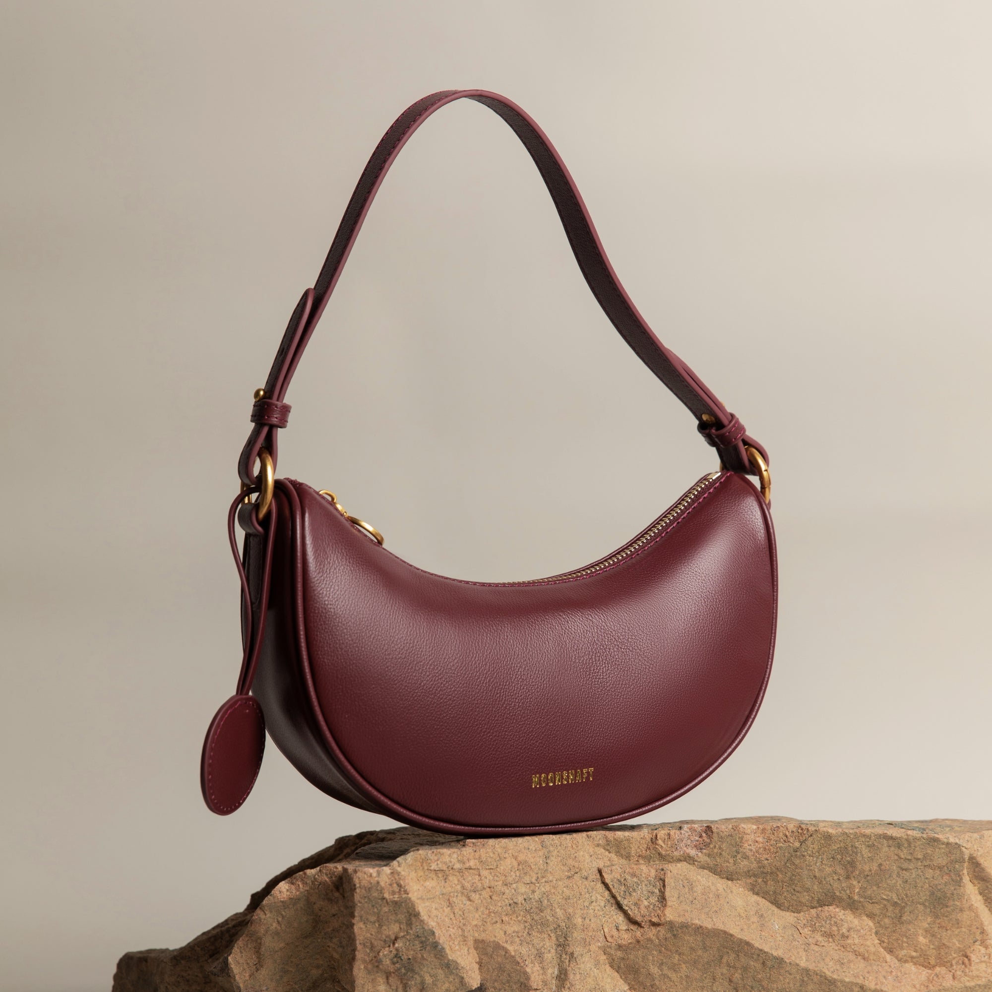 Crescent Moon bag in wine smooth leather, elegantly displayed on a stone surface with gold hardware details. The bag's curved silhouette and minimalist design are complemented by a detachable leather tag, set against a neutral-toned background, highlighting its premium craftsmanship and modern aesthetic.