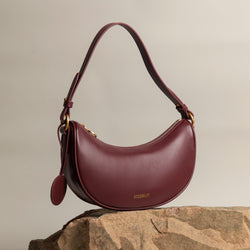 Crescent Moon bag in wine smooth leather, elegantly displayed on a stone surface with gold hardware details. The bag's curved silhouette and minimalist design are complemented by a detachable leather tag, set against a neutral-toned background, highlighting its premium craftsmanship and modern aesthetic.