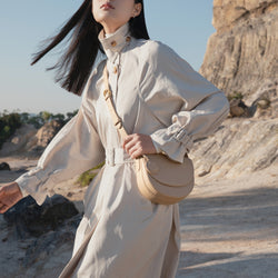 Model wearing a beige trench coat, holding a beige half moon crossbody bag against an outdoor rocky landscape, highlighting the minimalist and sophisticated design of the bag with a circular leather tag.