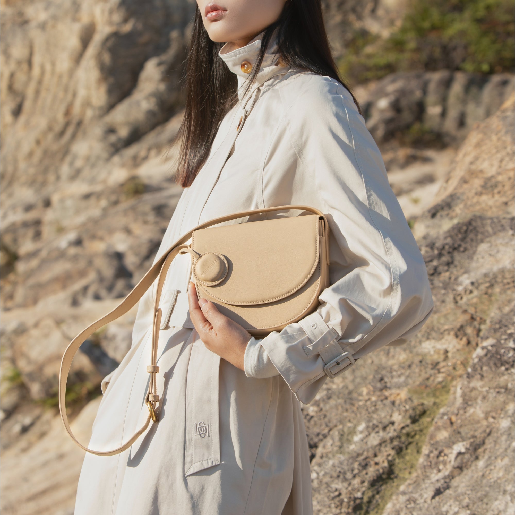 Model holding a beige half moon crossbody bag with a circular leather tag, dressed in a beige trench coat, against a rocky outdoor background, emphasizing the elegant and natural tones of the bag's design.