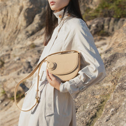 Model holding a beige half moon crossbody bag with a circular leather tag, dressed in a beige trench coat, against a rocky outdoor background, emphasizing the elegant and natural tones of the bag's design.