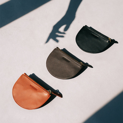 Three Half Moon Coin Purses in different colors—cognac, brown, and black—laid flat on a smooth surface. The purses are arranged diagonally, with a distinct shadow cast by a hand reaching toward them, creating a play of light and shadow that adds visual interest to the composition. Each purse has a gold zipper and an embossed MOONSHAFT logo.