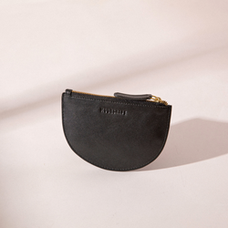 The image features the Half Moon Coin Purse in black goat leather, standing upright against a light background. The MOONSHAFT logo is embossed on the front, and the purse is equipped with a gold-tone zipper along the top, complemented by a matching black leather pull. Gentle shadows cast from the product emphasize its sleek design and soft leather texture.