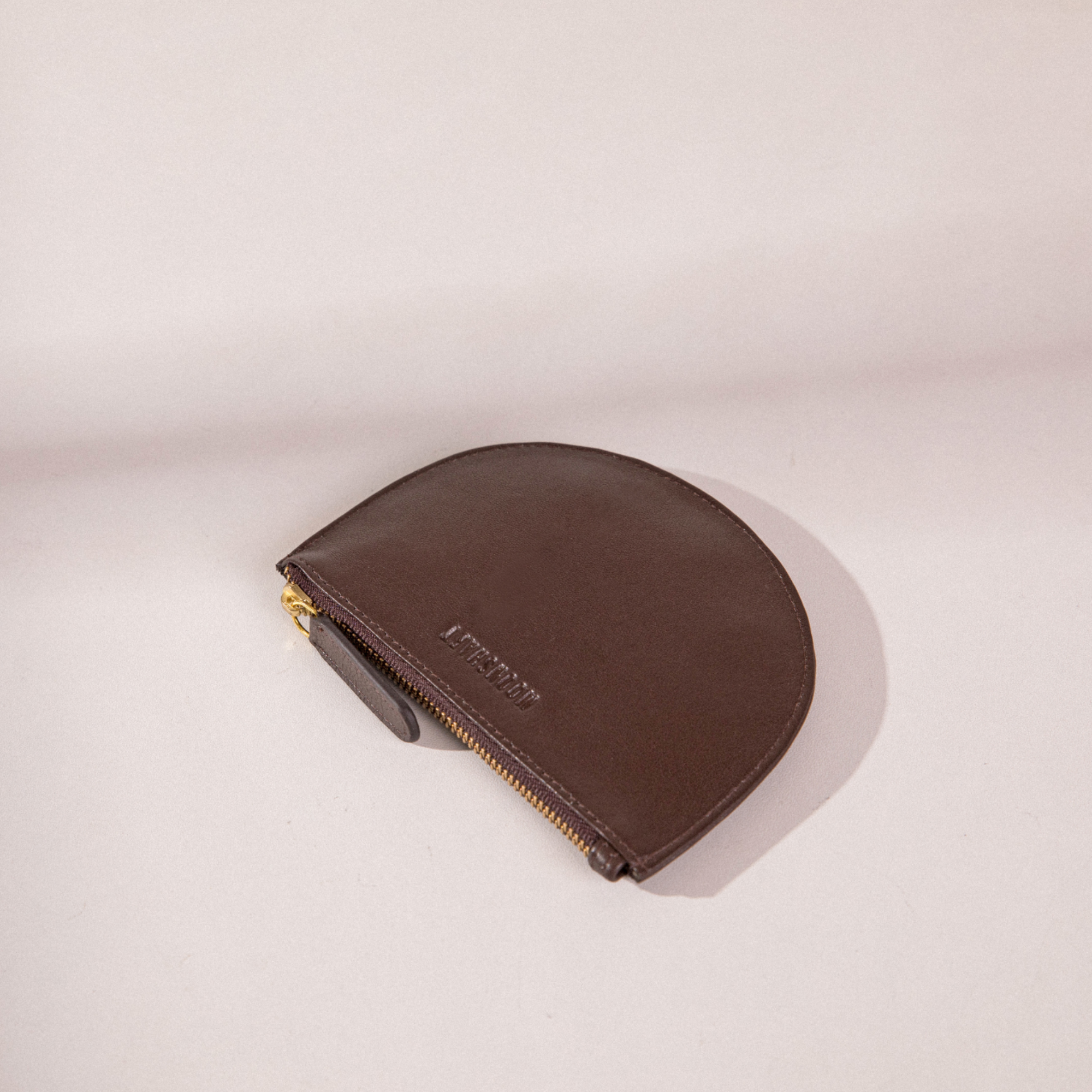 The image shows a brown Half Moon Coin Purse placed flat on a light background. The purse is made of goat leather, featuring the MOONSHAFT logo embossed on the front and a gold zipper with a matching leather pull tab. Shadows cast by the purse add depth to the simple yet elegant composition.