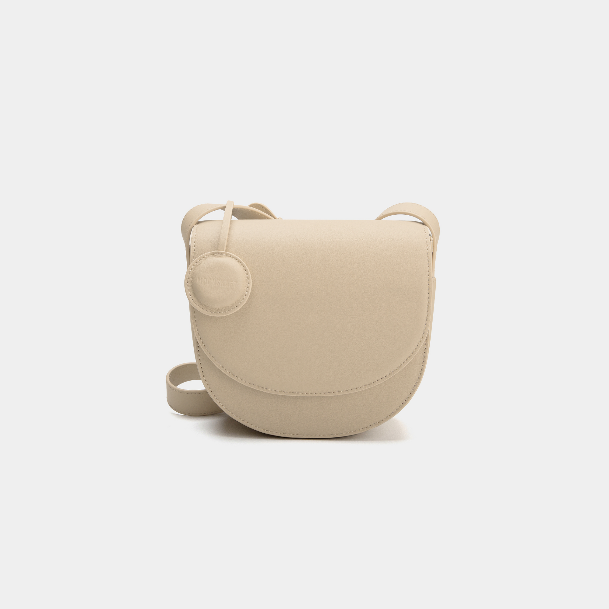 A beige half-moon crossbody bag with a smooth leather finish, featuring a circular leather tag attached to the strap. The structured design highlights the bag's minimalist aesthetic and sleek curves, offering an elegant yet functional style for everyday wear.