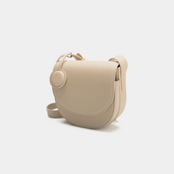 A front-side angle view of a beige half-moon crossbody bag made of smooth leather. The bag has a curved base and minimalistic flap closure. A circular leather tag is attached to the strap, adding a subtle design detail. The adjustable strap and sleek design offer a versatile, elegant look.