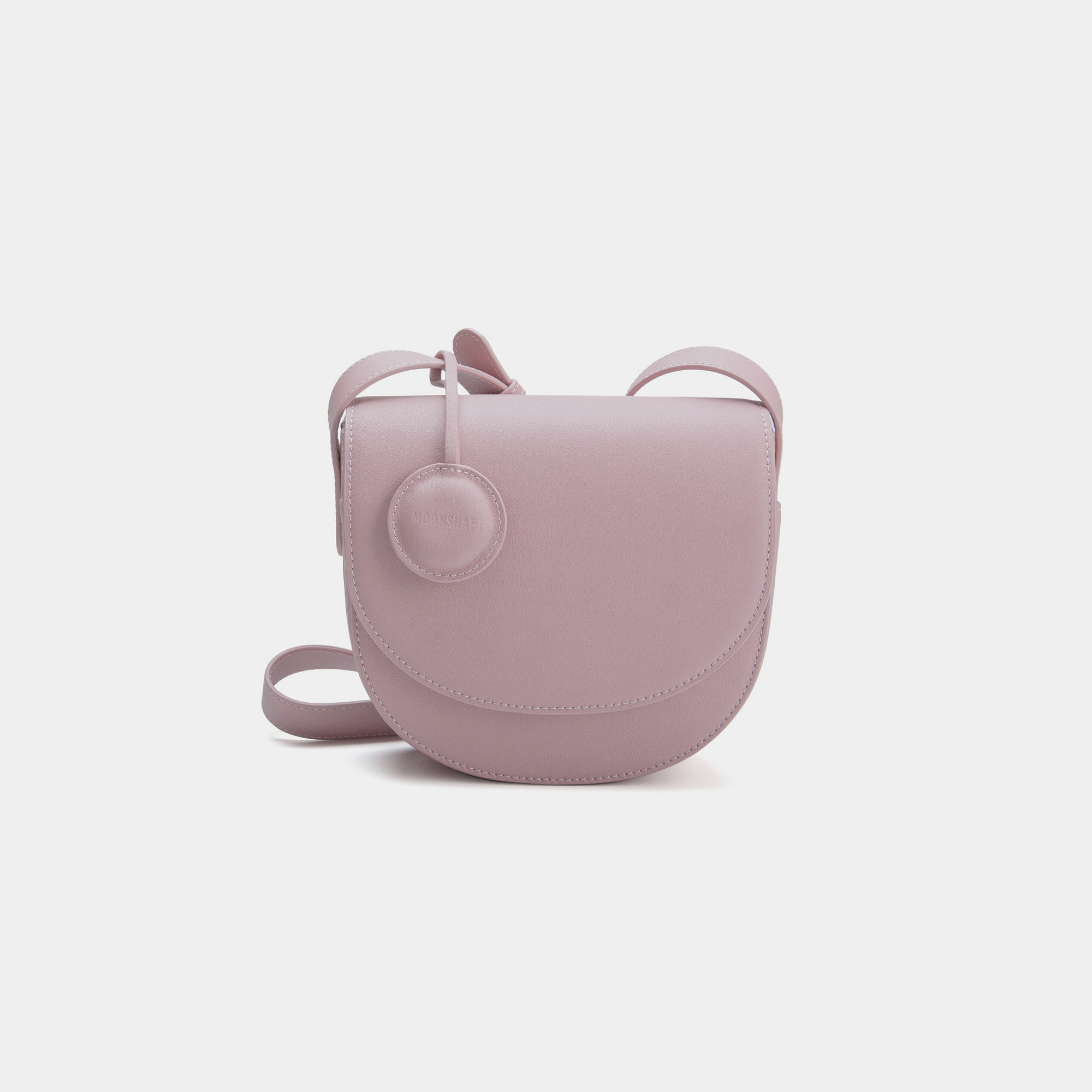 A Light Lilac half-moon crossbody bag with a sleek, smooth leather finish. It features a circular leather tag attached to the strap and a simple, structured design. The bag has a curved flap closure and an adjustable crossbody strap.
