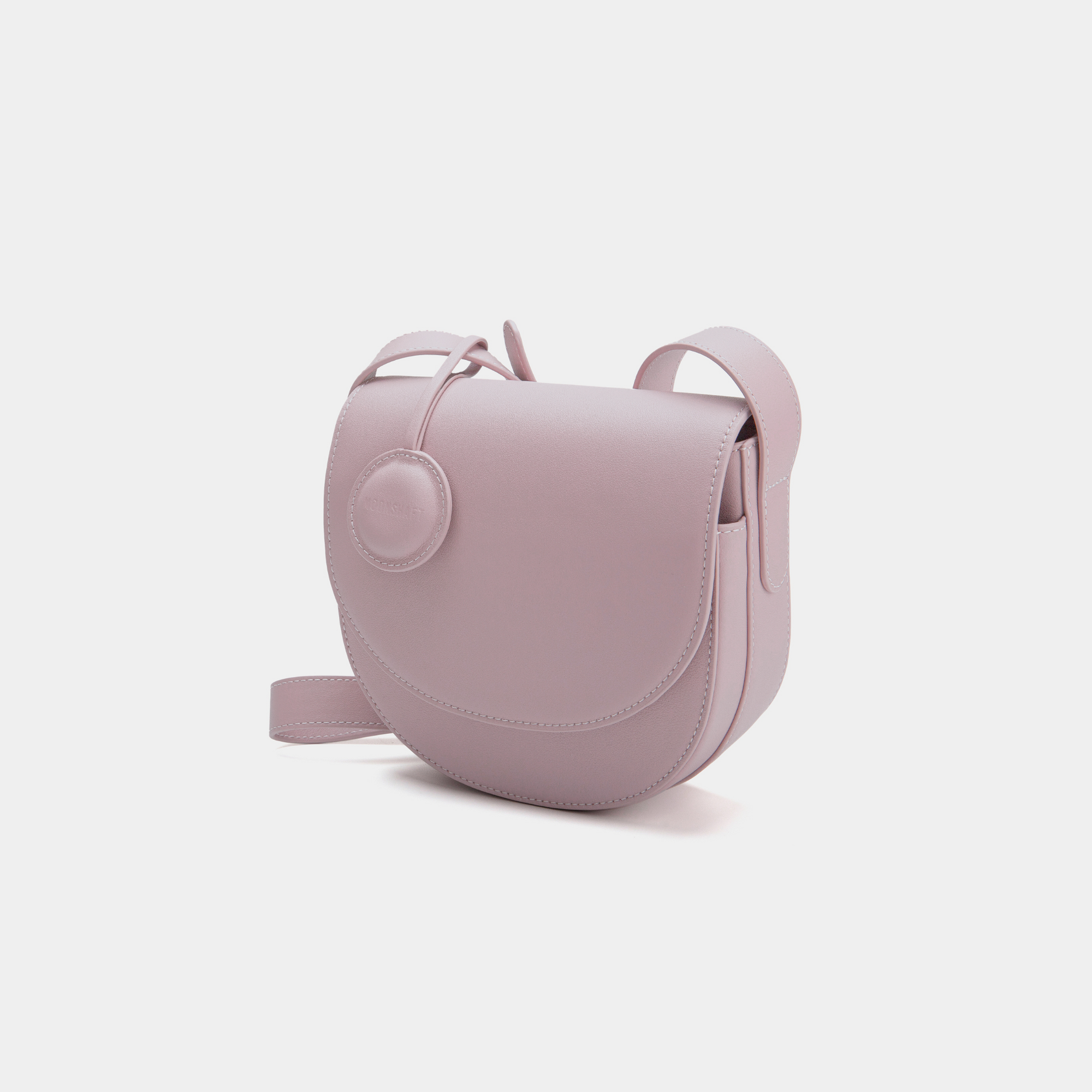 A front-side angle view of a light lilac half-moon crossbody bag made of smooth leather. The bag has a curved base and minimalistic flap closure. A circular leather tag is attached to the strap, adding a subtle design detail. The adjustable strap and sleek design offer a versatile, elegant look.