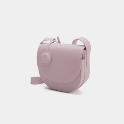A front-side angle view of a light lilac half-moon crossbody bag made of smooth leather. The bag has a curved base and minimalistic flap closure. A circular leather tag is attached to the strap, adding a subtle design detail. The adjustable strap and sleek design offer a versatile, elegant look.