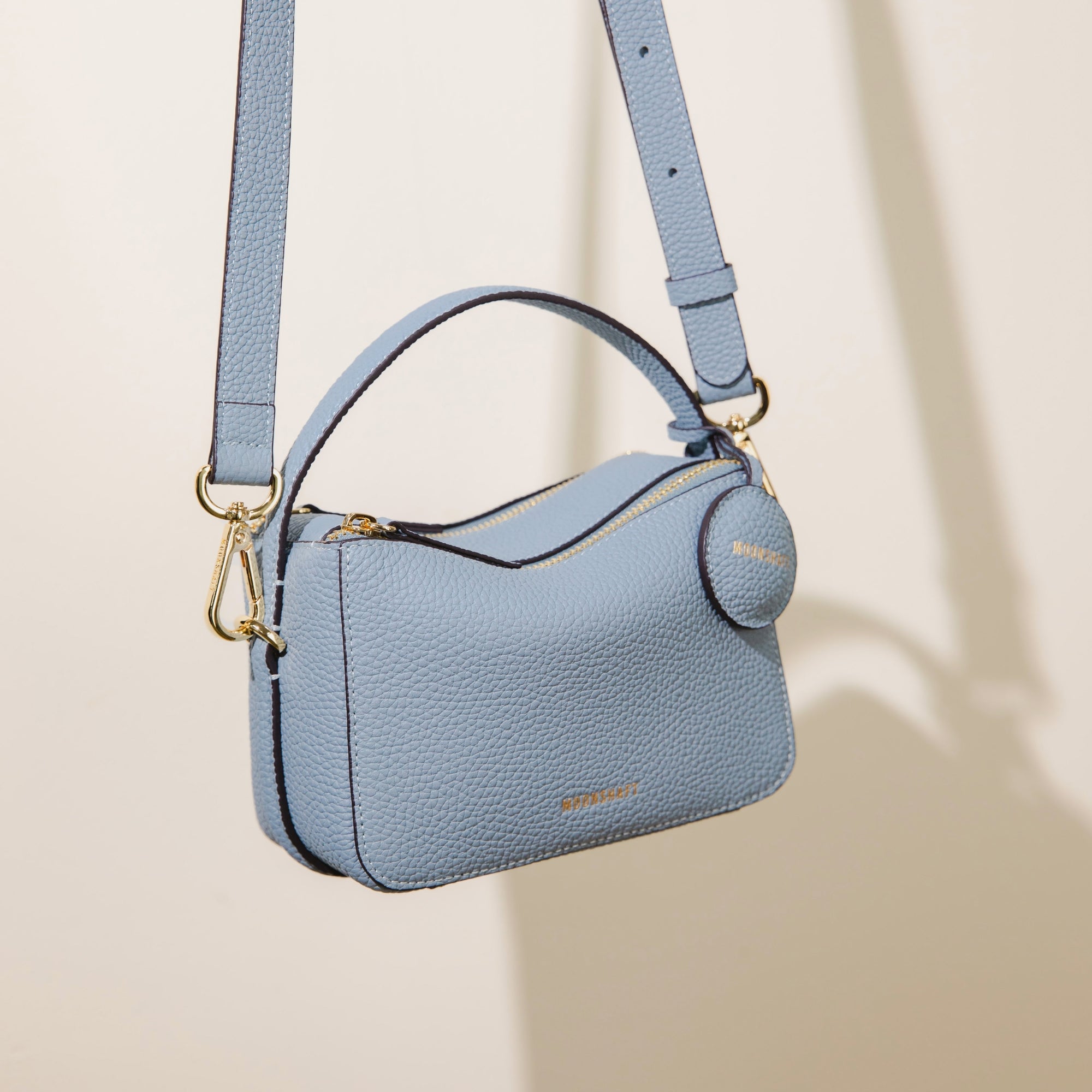The Mini Double Zip Bag in aquamarine color is shown hanging by its adjustable shoulder strap. The bag is made from pebbled leather, featuring a top handle, gold-tone hardware, and a round leather tag embossed with the 'MOONSHAFT' logo in gold. The clean, structured design highlights the bag's elegant and versatile style.