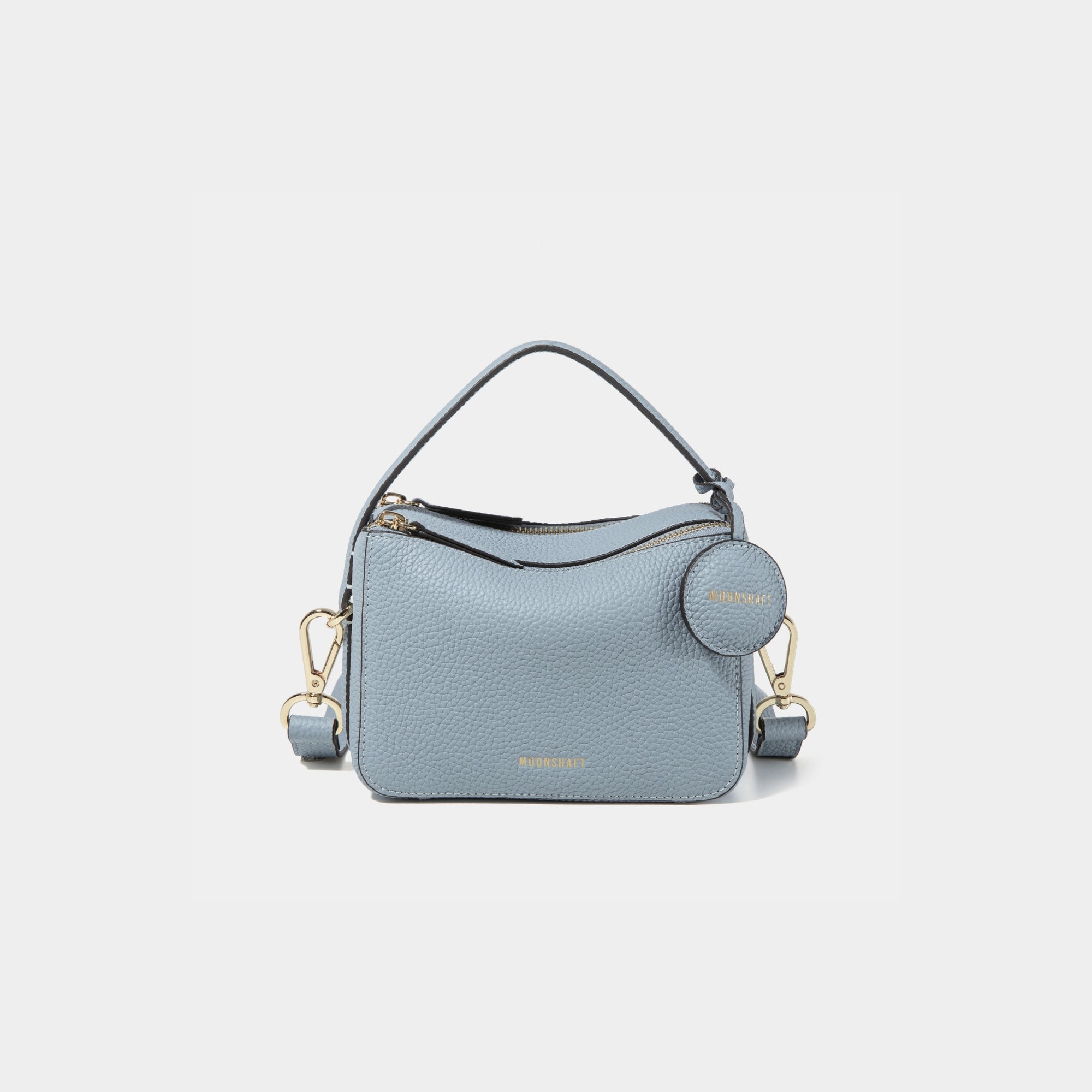 A close-up of the Mini Double Zip Bag in a soft blue shade, made from pebbled leather. The bag features a top handle and an adjustable shoulder strap with gold-tone hardware. It has two zippered compartments for storage and a round leather tag attached to the side, embossed with the brand logo 'MOONSHAFT' in gold. The minimalist and structured design enhances its modern and stylish appeal.