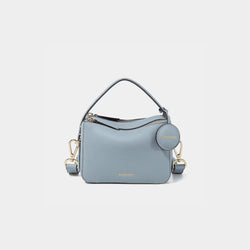 A close-up of the Mini Double Zip Bag in a soft blue shade, made from pebbled leather. The bag features a top handle and an adjustable shoulder strap with gold-tone hardware. It has two zippered compartments for storage and a round leather tag attached to the side, embossed with the brand logo 'MOONSHAFT' in gold. The minimalist and structured design enhances its modern and stylish appeal.