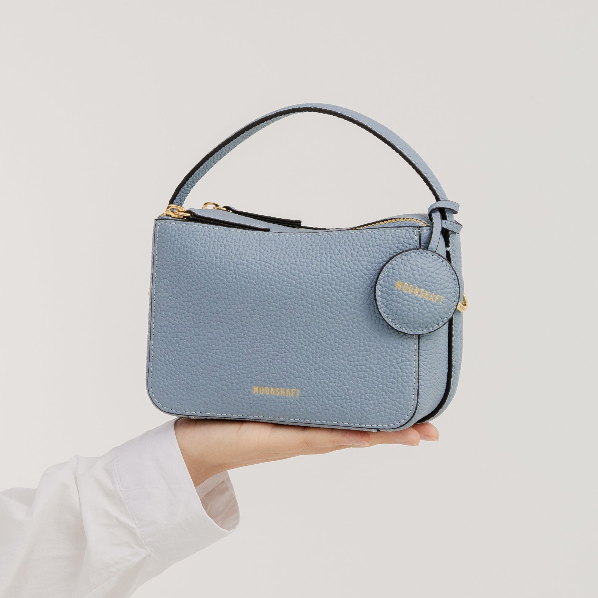 The Mini Double Zip Bag in aquamarine color is held in a hand, showcasing its compact, structured design. Made from pebbled leather, the bag features a top handle, gold-tone hardware, and a round leather tag embossed with the 'MOONSHAFT' logo in gold. The clean and minimalist design highlights the bag's elegant and functional style.