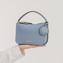 The Mini Double Zip Bag in aquamarine color is held in a hand, showcasing its compact, structured design. Made from pebbled leather, the bag features a top handle, gold-tone hardware, and a round leather tag embossed with the 'MOONSHAFT' logo in gold. The clean and minimalist design highlights the bag's elegant and functional style.