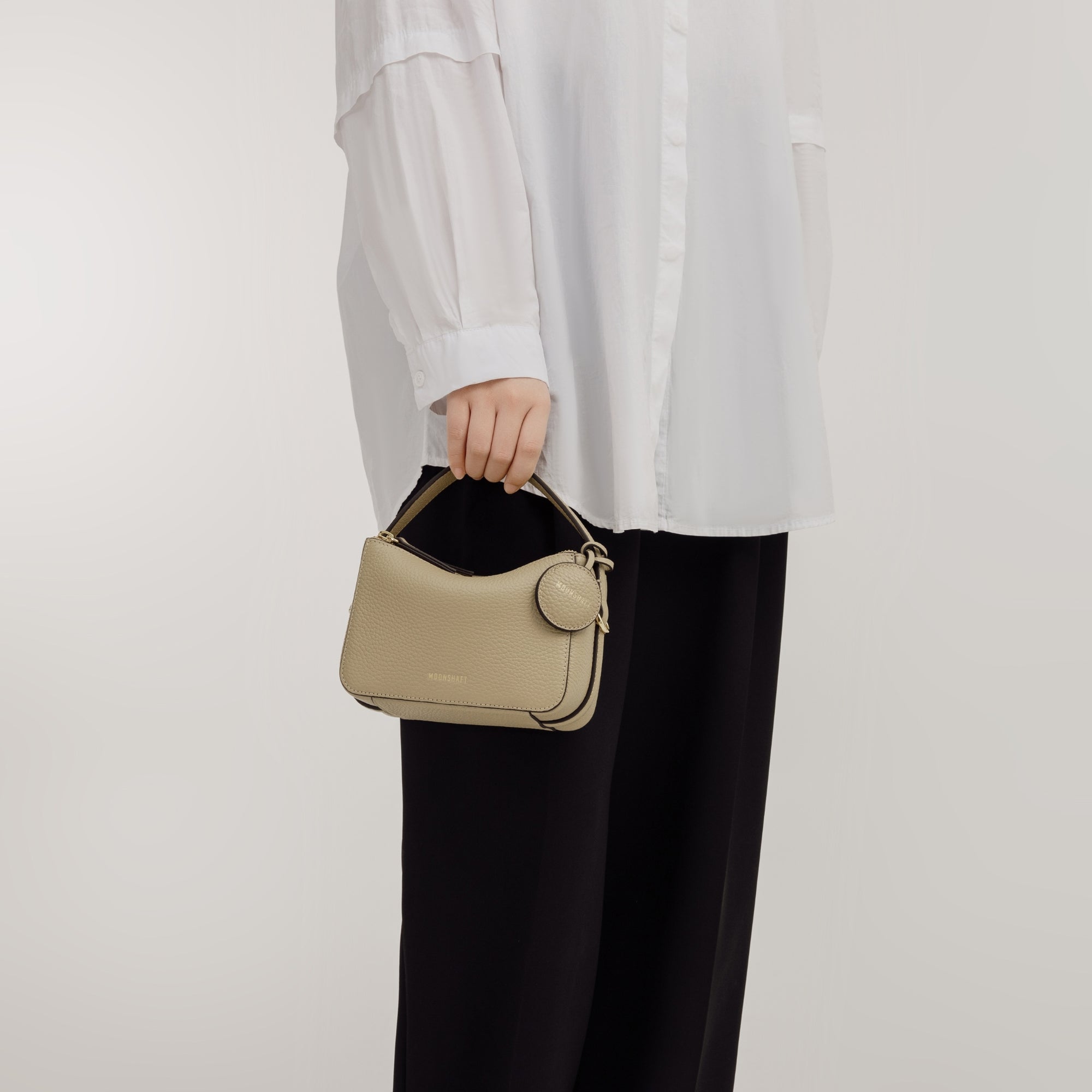 A model in a white oversized shirt and black pants holds the Mini Double Zip Bag in a neutral beige color by its top handle. The bag is crafted from pebbled leather with a structured silhouette, featuring a round leather tag and gold-tone hardware. The minimalist design complements the model's casual, chic outfit, showcasing the bag's elegance and versatility.