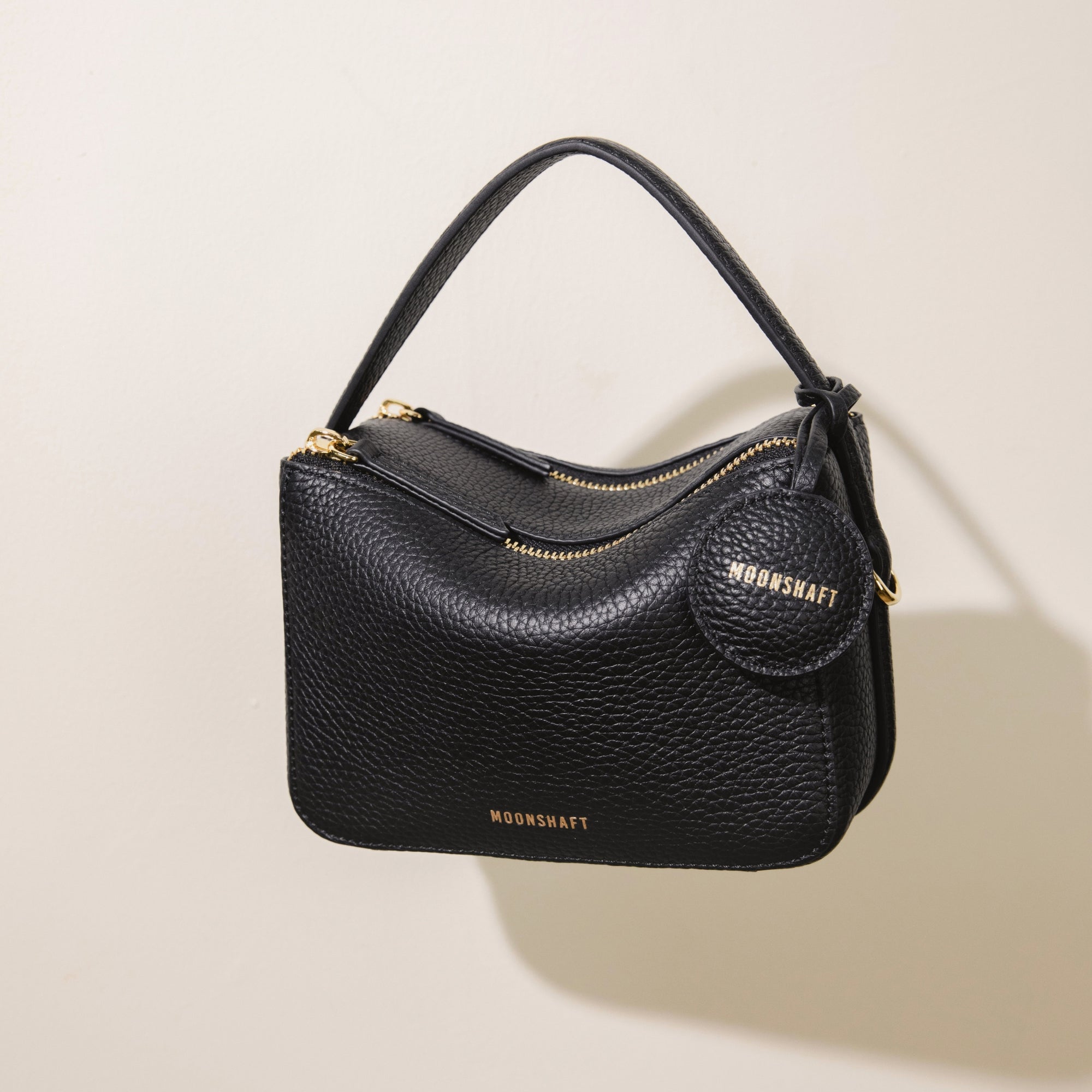 The Mini Double Zip Bag in black, made from pebbled leather, is shown hanging by its top handle. The bag features gold-tone hardware, including two zippered compartments and a round leather tag embossed with the 'MOONSHAFT' logo. The structured design and minimalist aesthetic highlight the bag's timeless and sophisticated style.