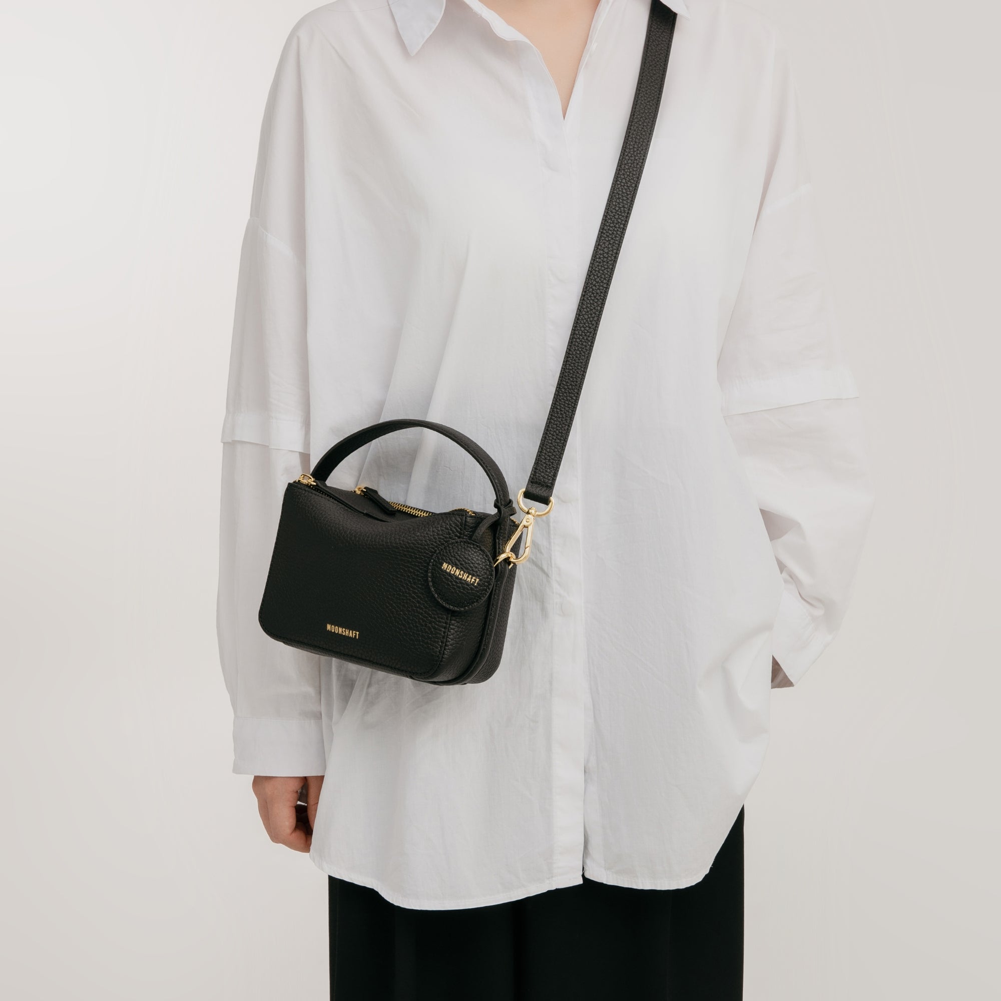 A model wearing a white oversized shirt and black pants carries the Mini Double Zip Bag in black, made from pebbled leather. The bag is styled crossbody with a gold-tone adjustable strap and features a top handle. The structured design is complemented by a round leather tag and gold-tone hardware, enhancing the bag's chic and minimalist appeal.