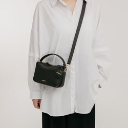 A model wearing a white oversized shirt and black pants carries the Mini Double Zip Bag in black, made from pebbled leather. The bag is styled crossbody with a gold-tone adjustable strap and features a top handle. The structured design is complemented by a round leather tag and gold-tone hardware, enhancing the bag's chic and minimalist appeal.