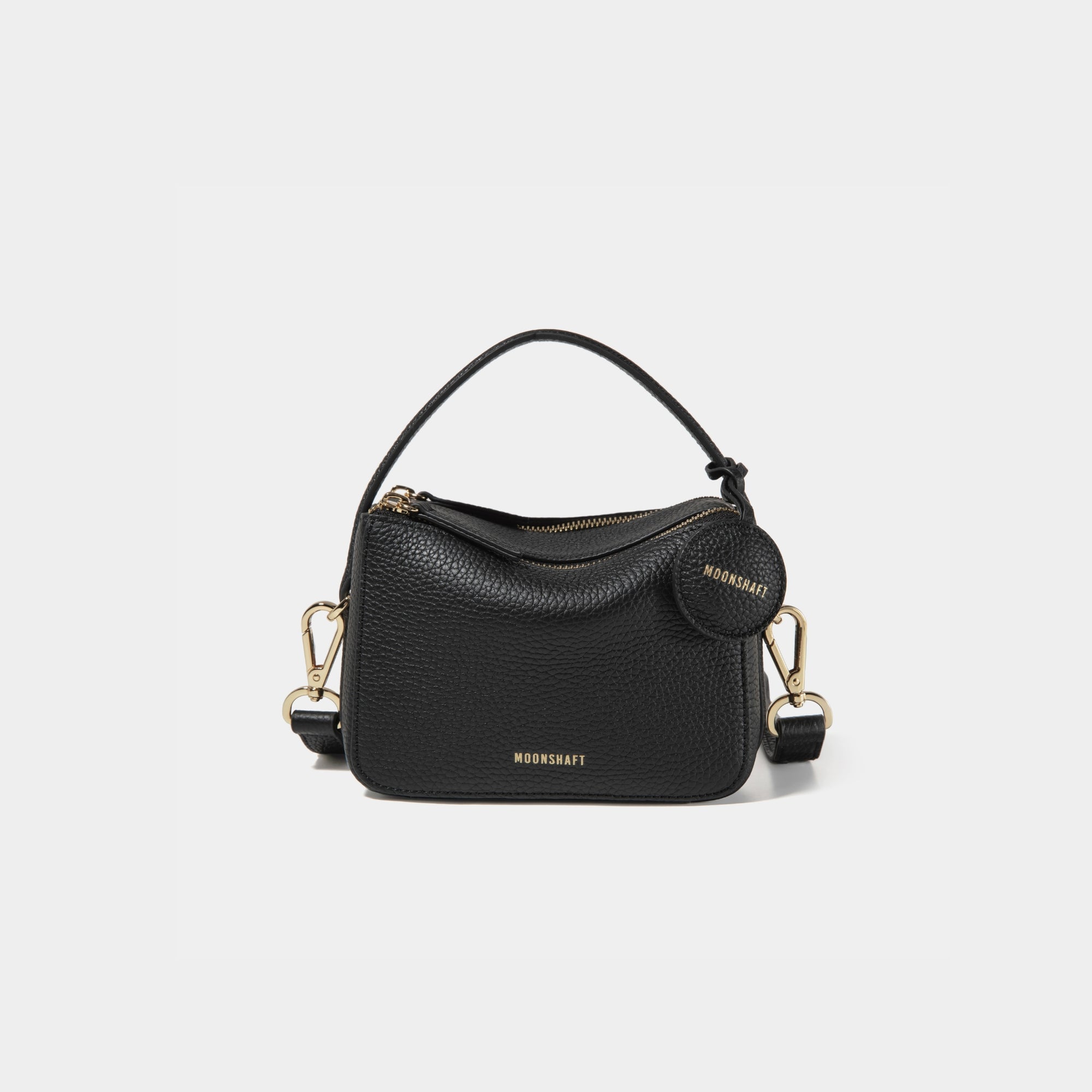 A close-up of the Mini Double Zip Bag in black color, made from pebbled leather. The bag features a top handle and an adjustable shoulder strap with gold-tone hardware. It has two zippered compartments for storage and a round leather tag attached to the side, embossed with the brand logo 'MOONSHAFT' in gold. The minimalist and structured design enhances its modern and stylish appeal.