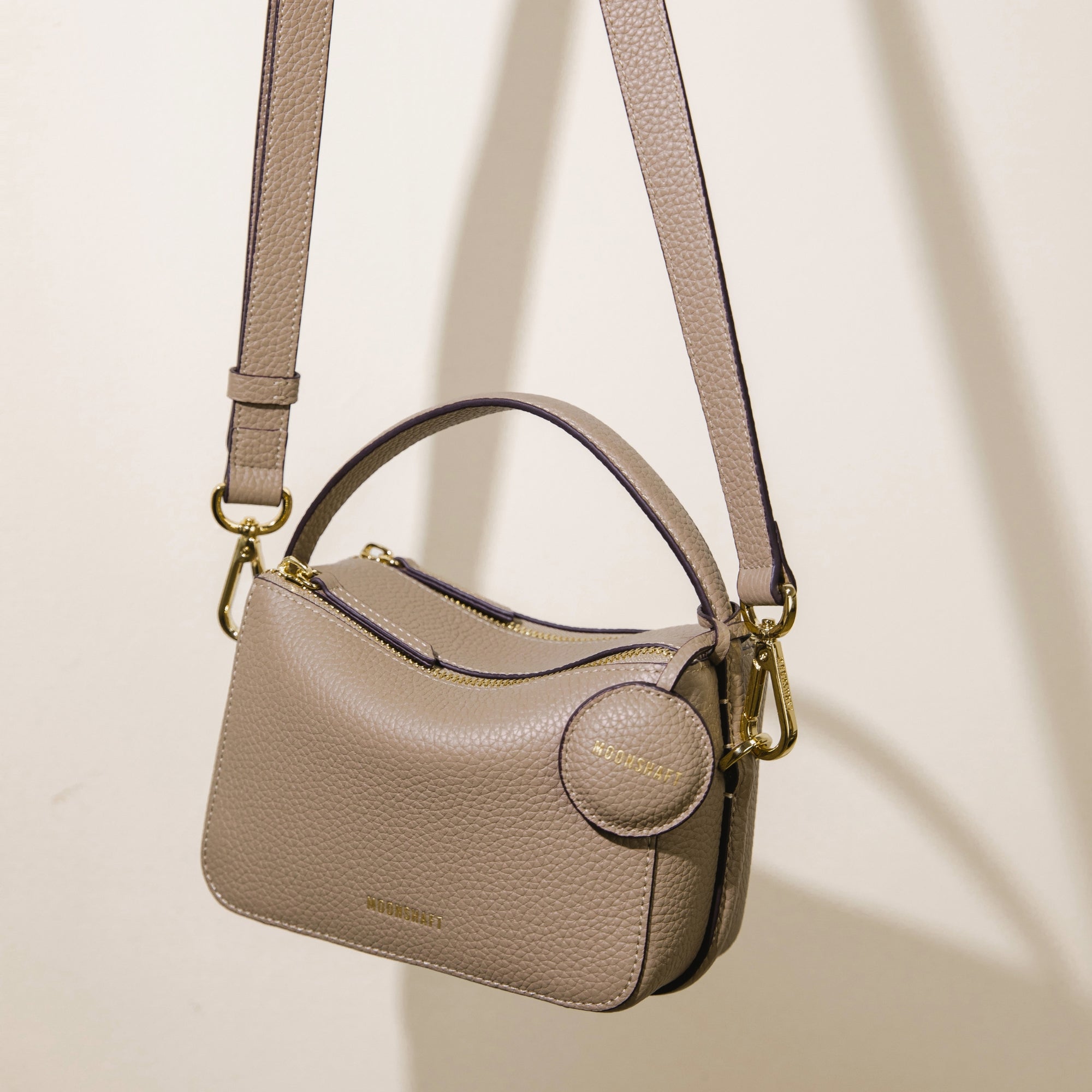 The Mini Double Zip Bag in sandstone color is shown hanging by its adjustable shoulder strap. The bag, made from pebbled leather, features a top handle, gold-tone hardware, and a round leather tag embossed with the 'MOONSHAFT' logo. The structured design and minimalist aesthetic highlight the bag's sophisticated and versatile style.